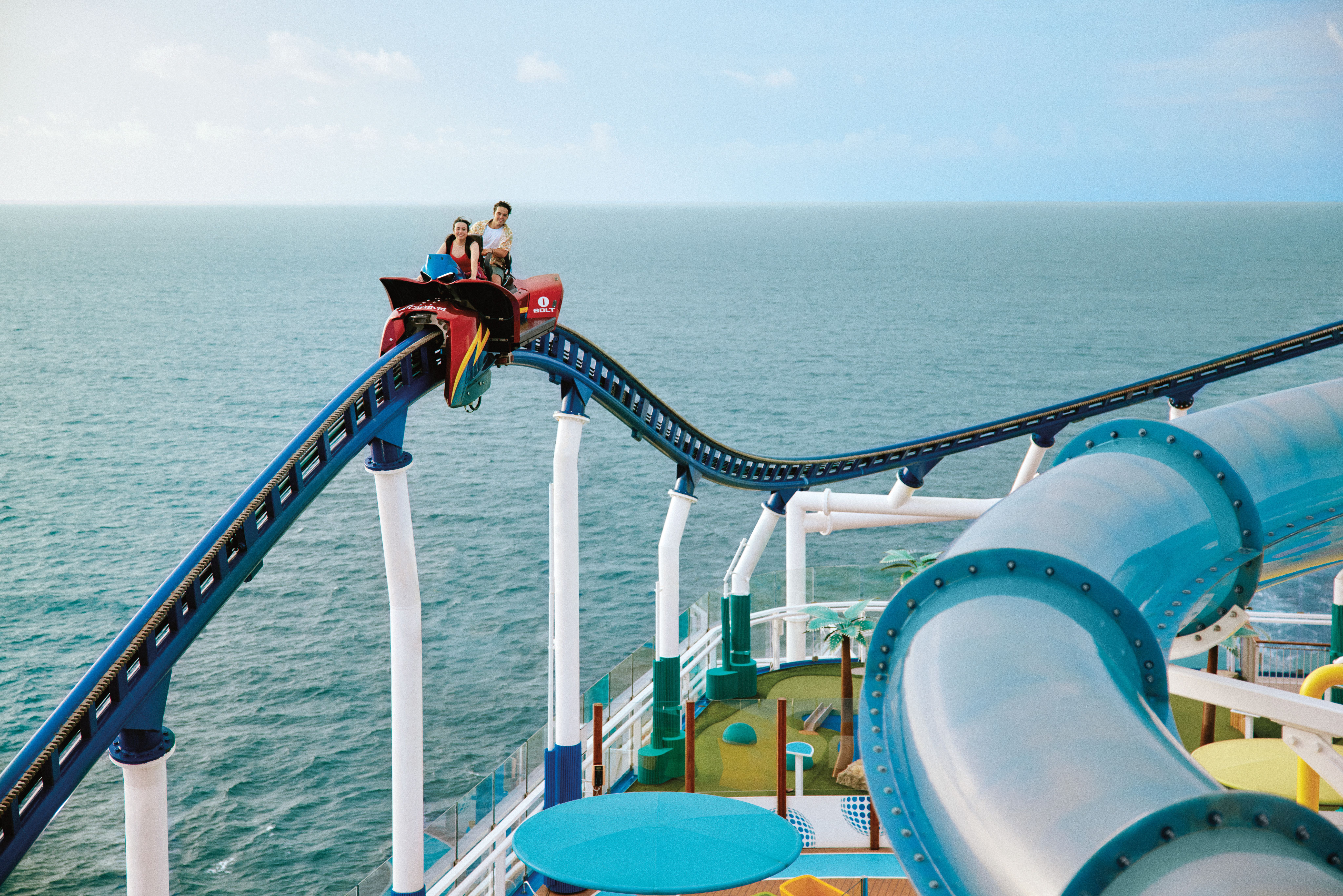 Couple riding Bolt roller coaster on Carnival Mardi Gras, soaring high above the sea with water slides and mini golf visible on deck.
