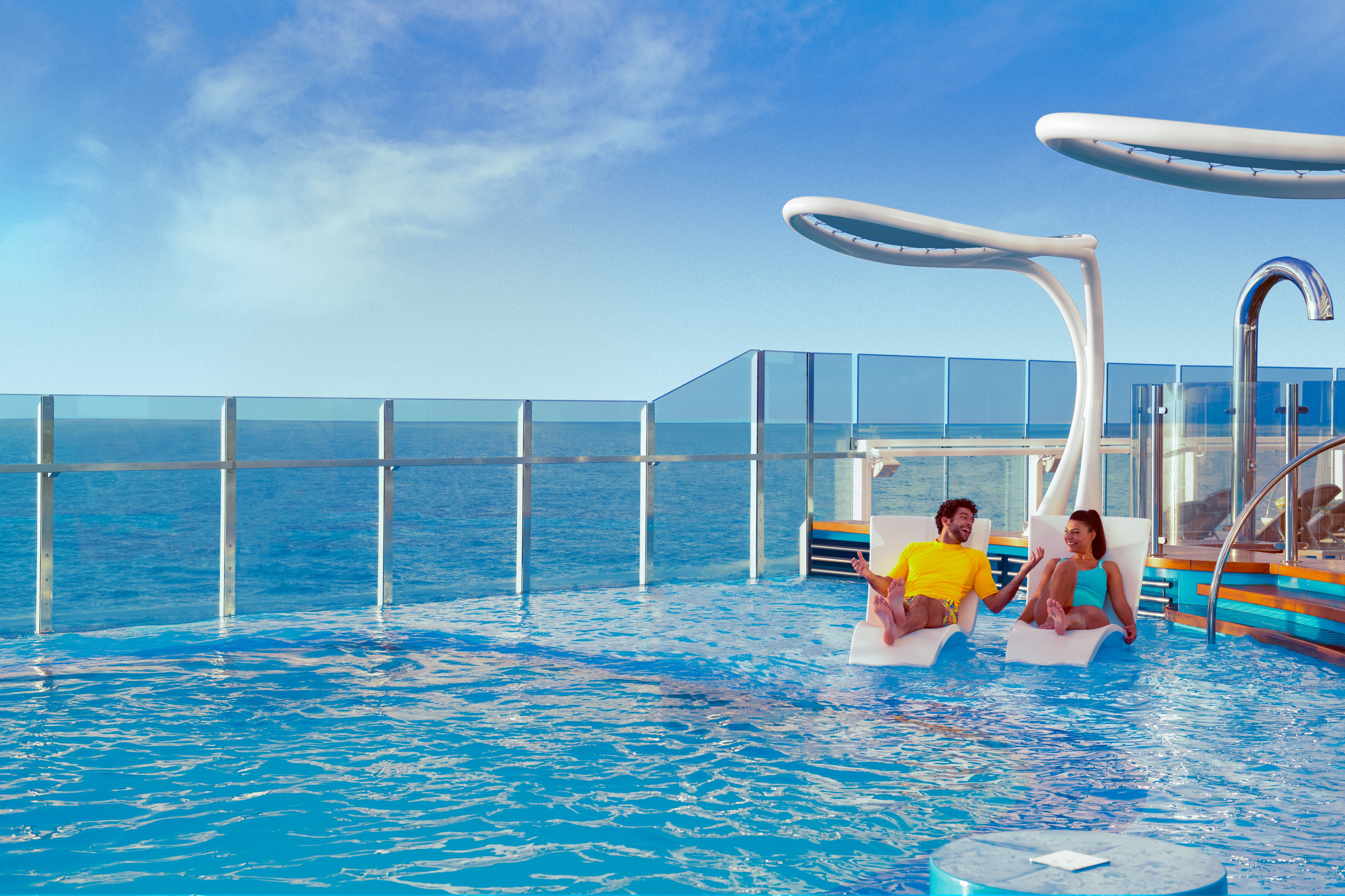 Couple relaxing on loungers in the Summer Landing infinity pool on Carnival Mardi Gras, with ocean views and modern design elements under sunny skies.
