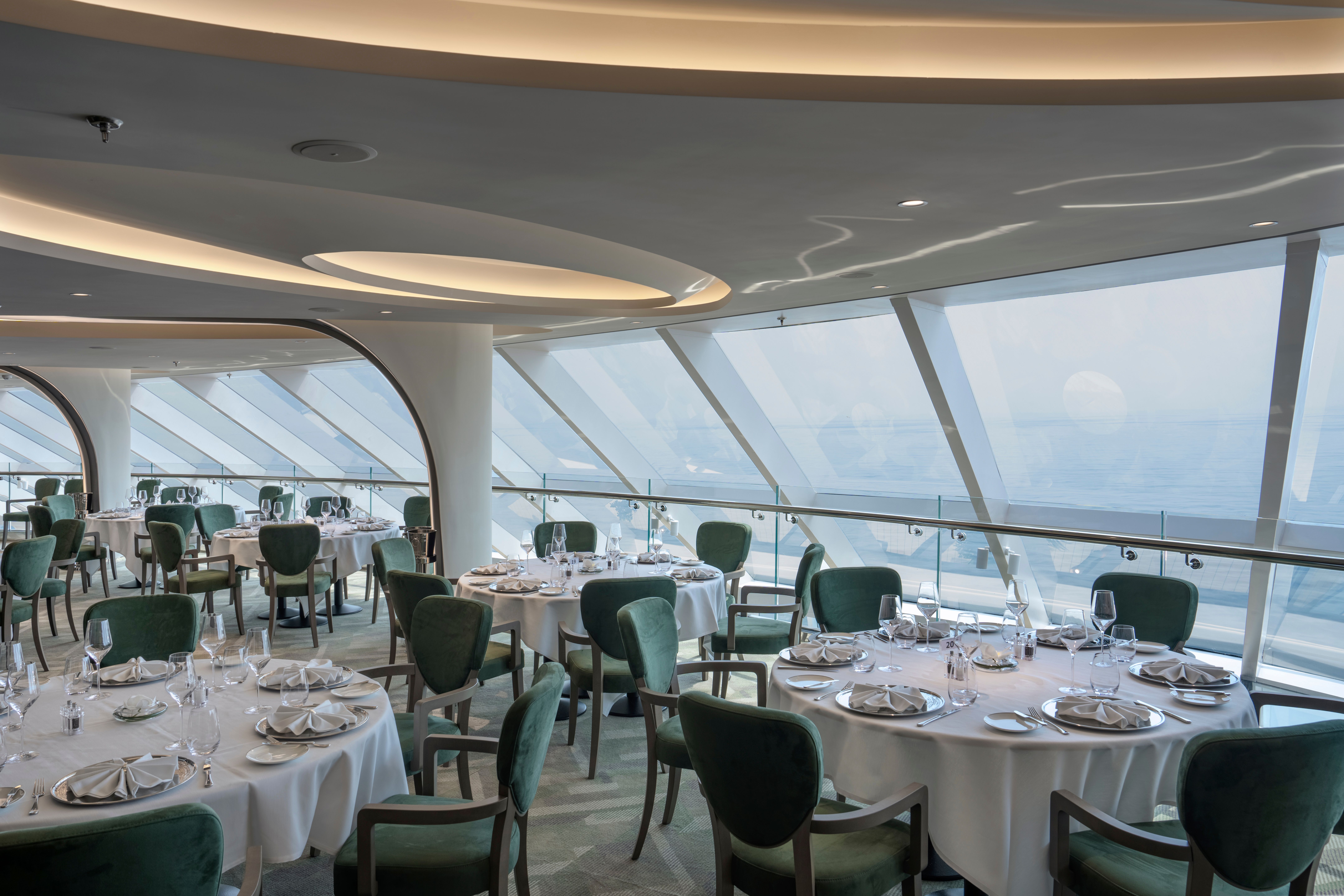 Elegant MSC Yacht Club restaurant with floor-to-ceiling ocean views, white tablecloth service, and plush green seating in a refined, panoramic setting.