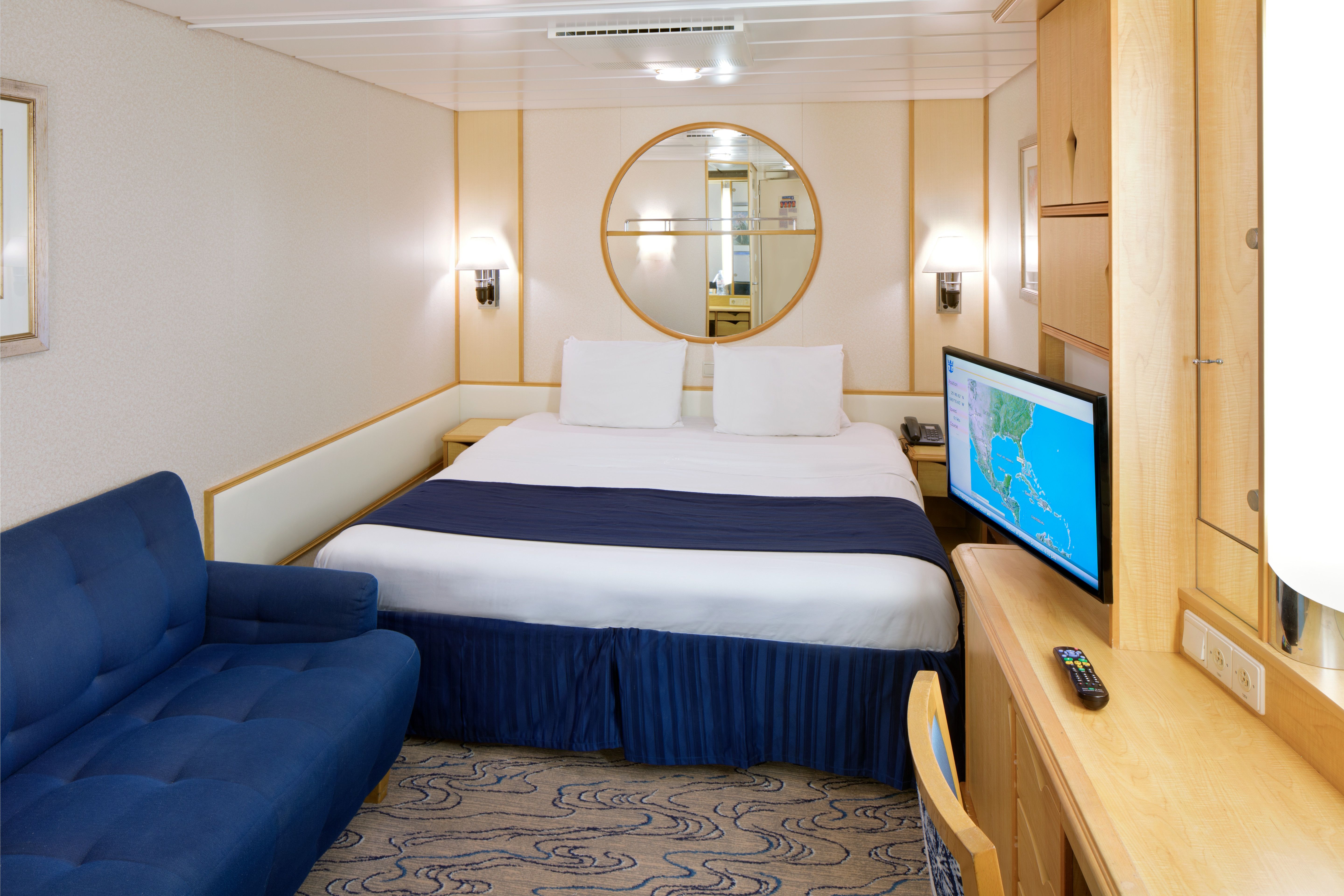Cozy interior stateroom on Royal Caribbean featuring a king bed, flat-screen TV, navy sofa, vanity desk, and mirror with warm wood accents.