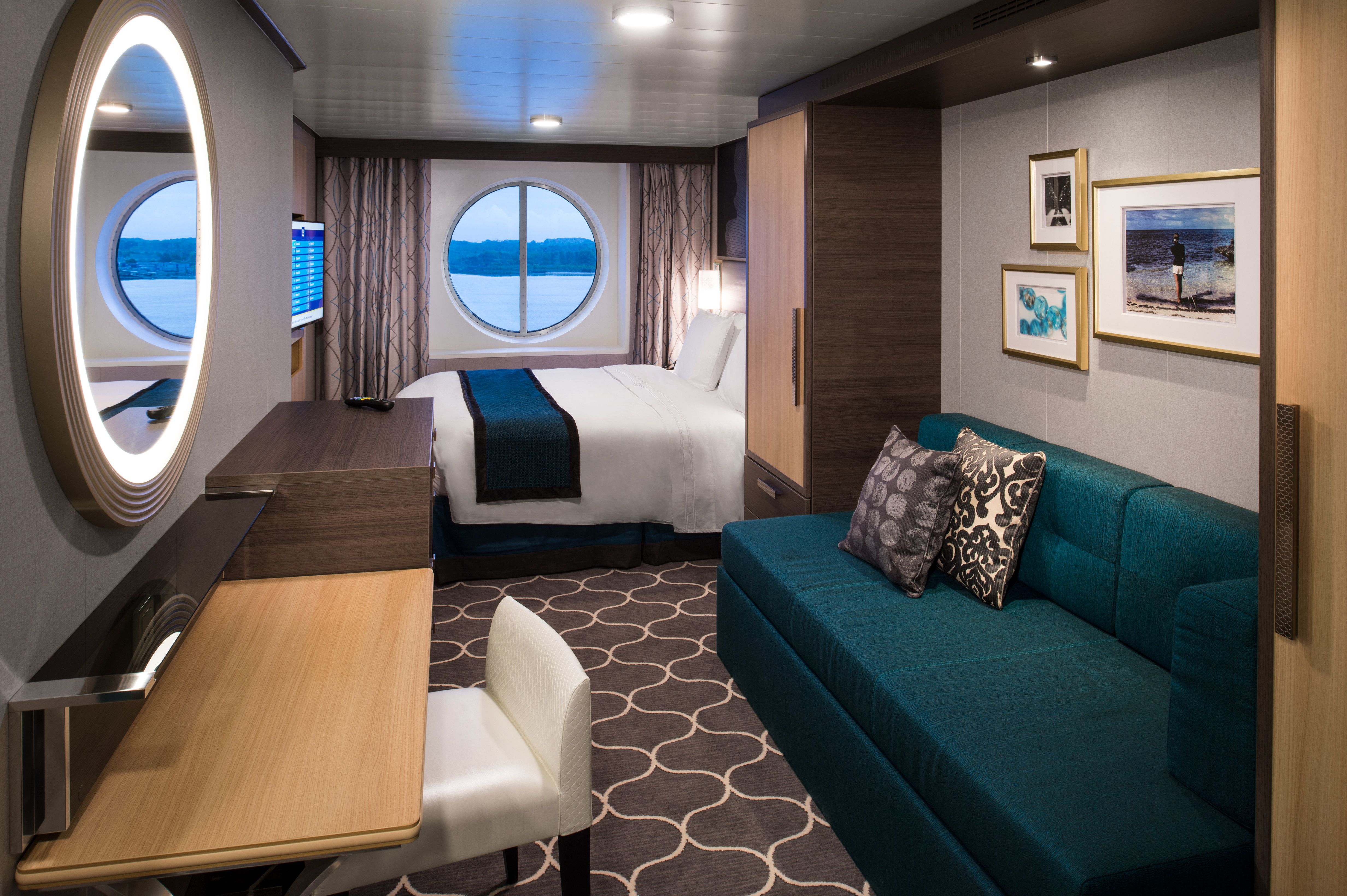 Stylish ocean view stateroom on Royal Caribbean featuring a round window, queen bed, teal sofa, modern desk, and elegant wall décor.