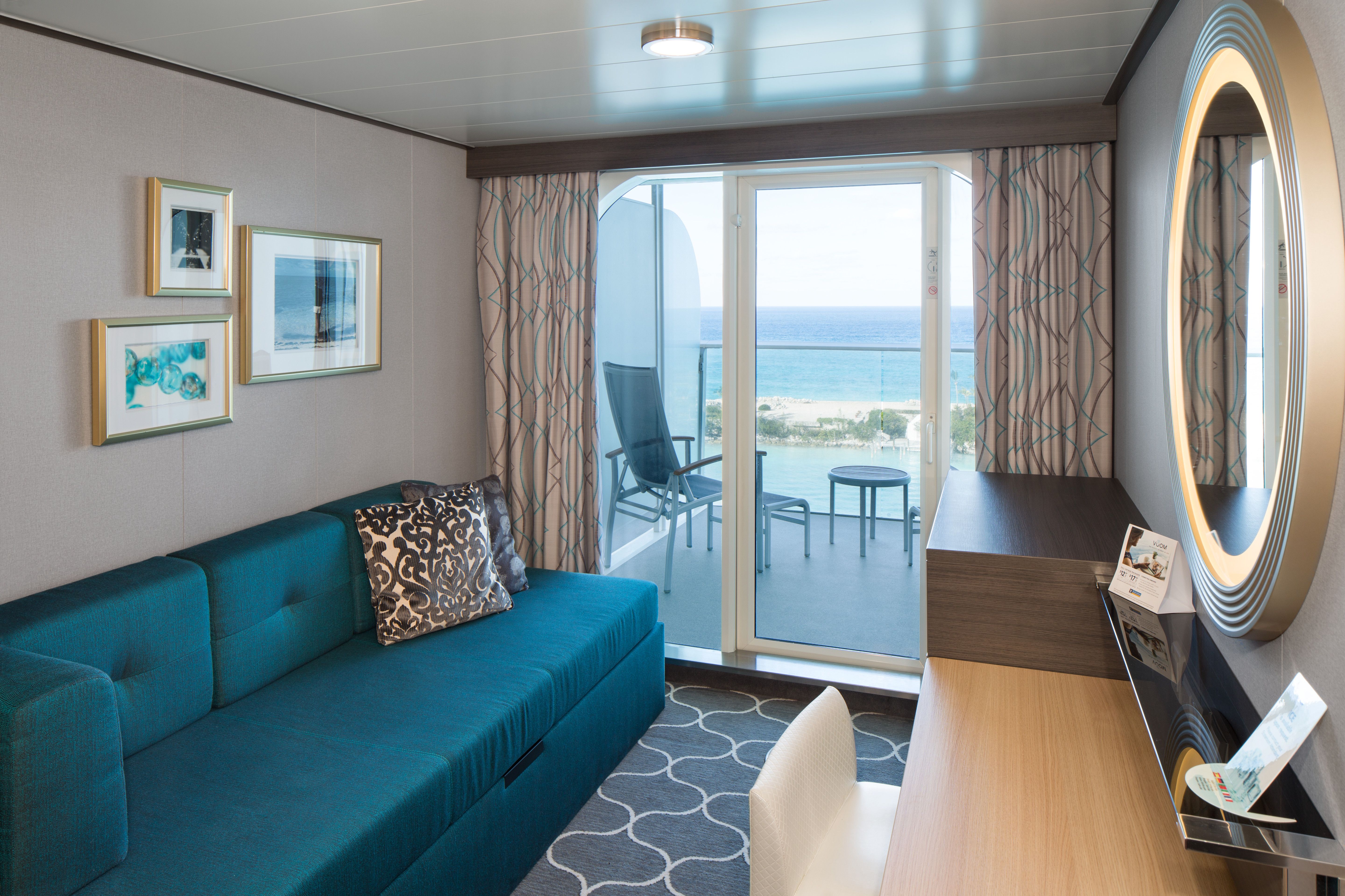 Modern Royal Caribbean balcony stateroom with teal sofa, vanity desk, and private furnished balcony overlooking the ocean and beach port.