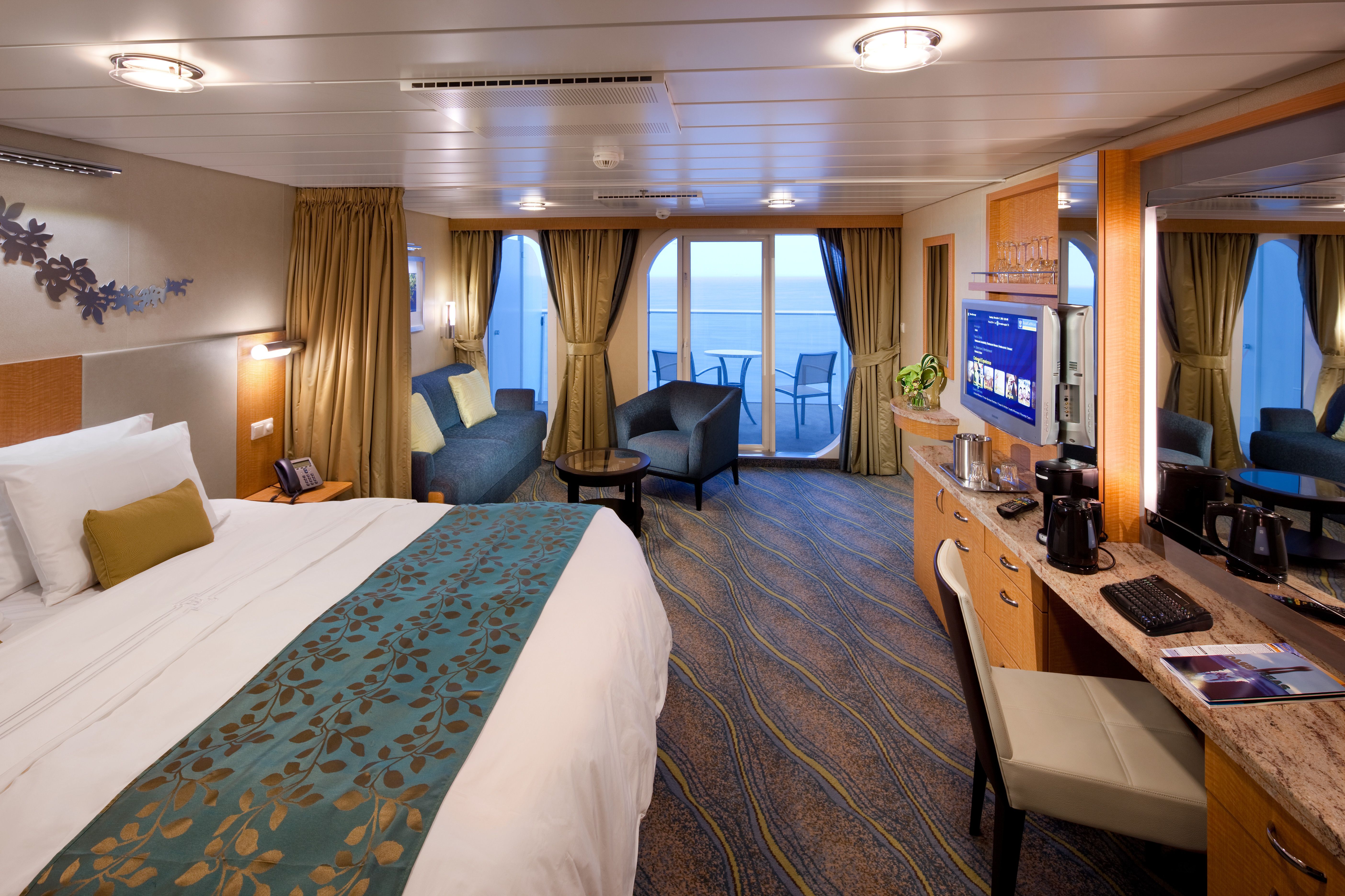 Spacious Royal Caribbean Junior Suite featuring a king bed, sitting area, desk, large balcony, and warm modern decor with ocean views.