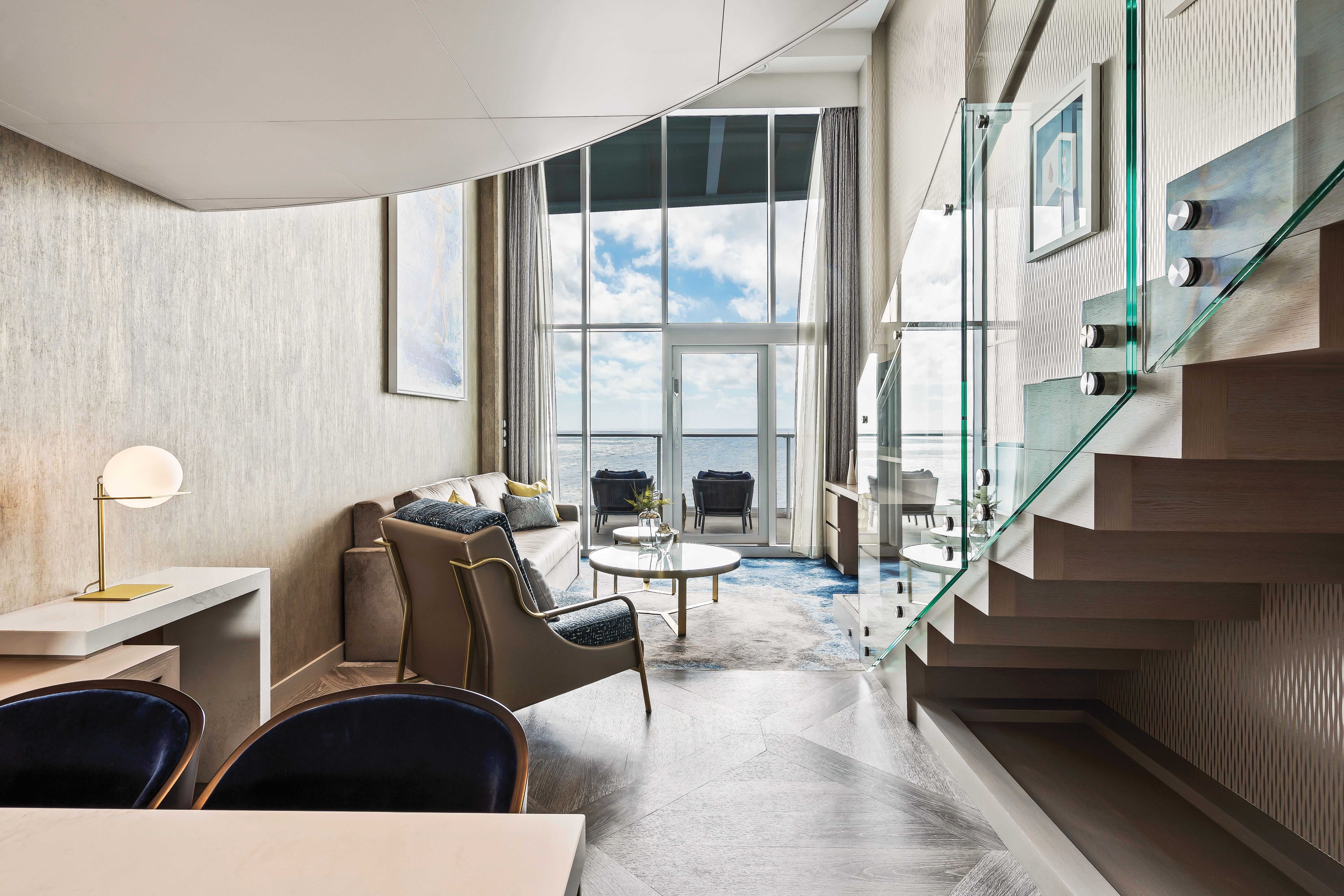 Elegant two-level Crown Loft Suite on Royal Caribbean with floor-to-ceiling windows, private balcony, glass staircase, and modern luxury decor.