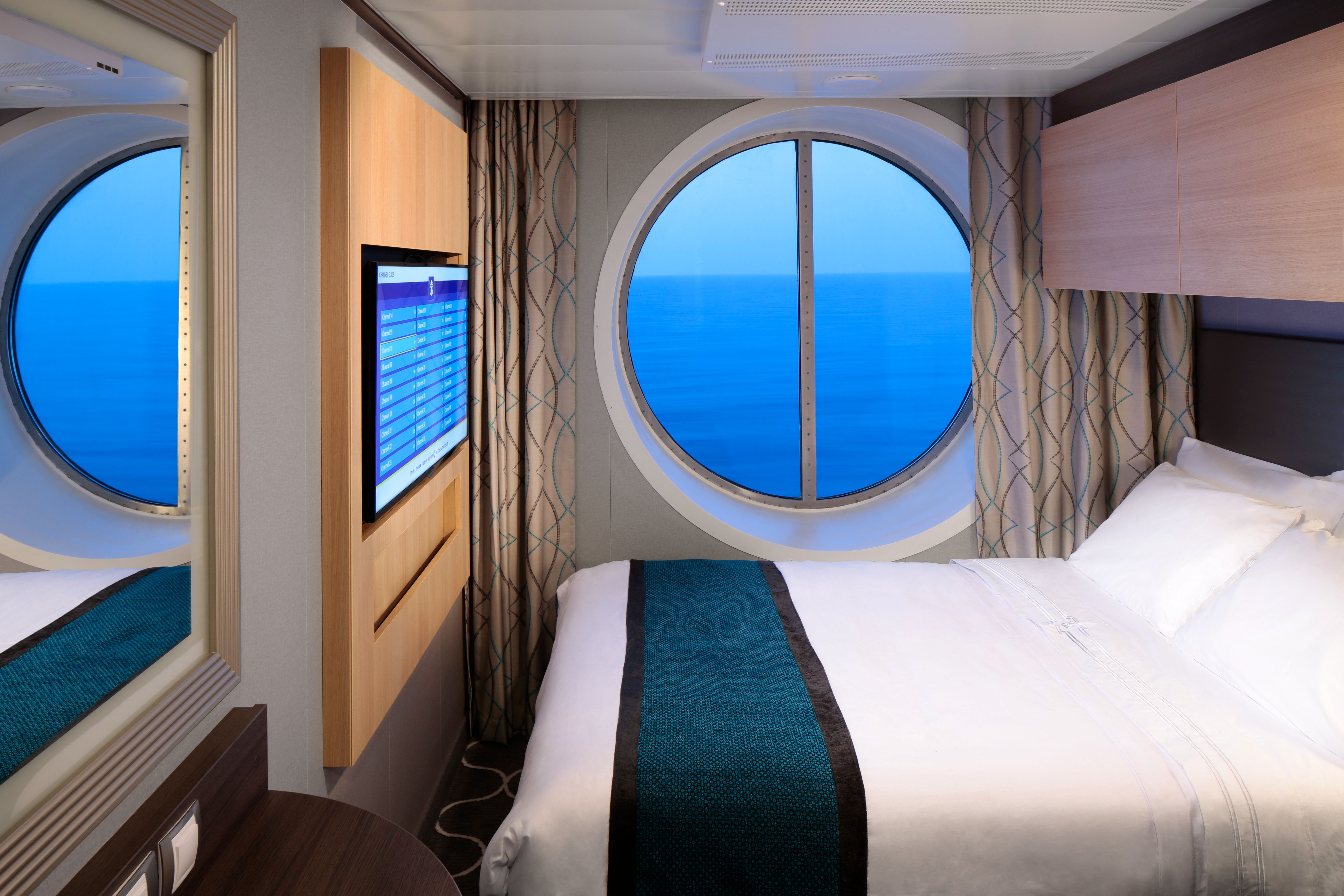 Compact studio cabin on Royal Caribbean with a large round porthole, queen bed, wall-mounted TV, and modern decor perfect for solo travelers.