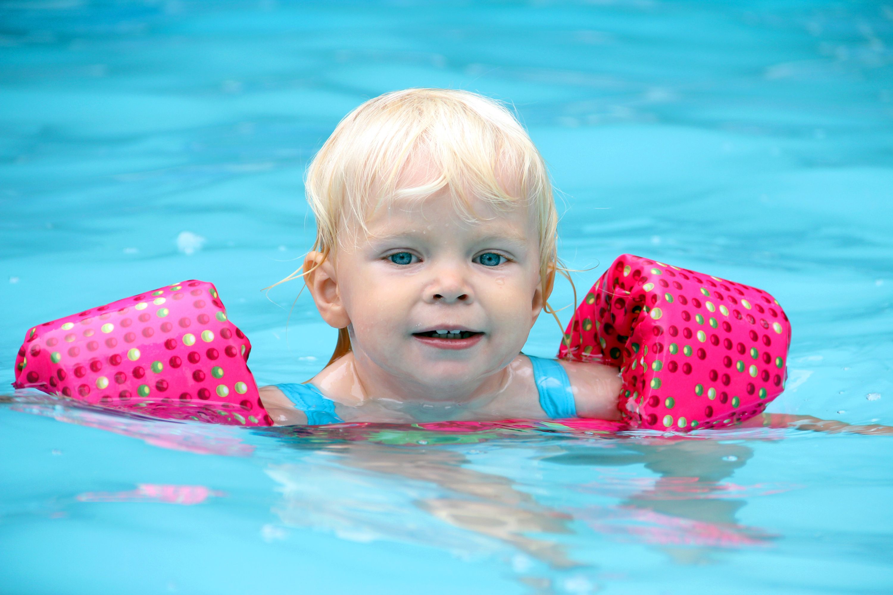 Blonde toddler enjoying a swim in a pool wearing pink polka-dot arm floaties, smiling confidently during water play.