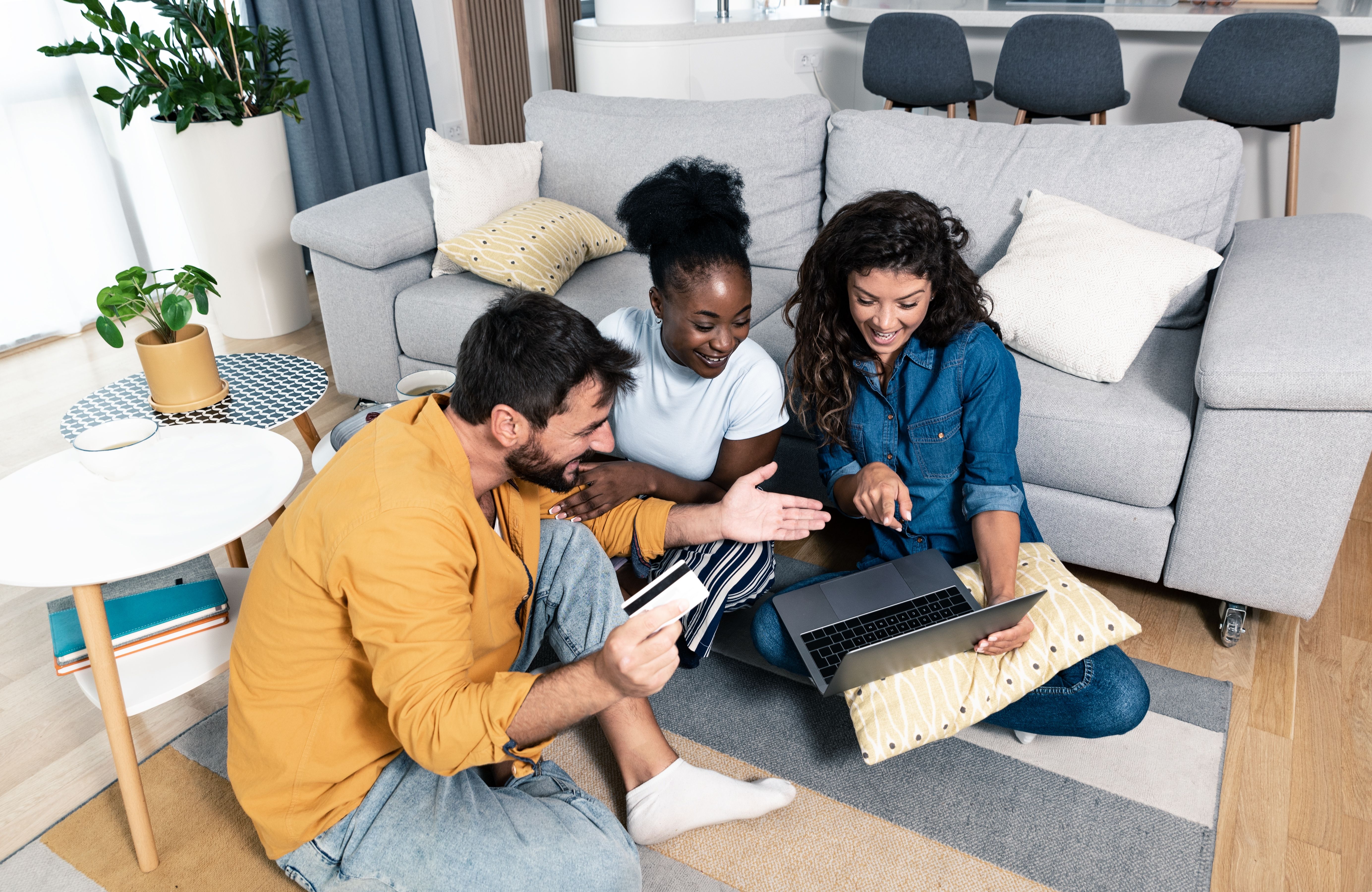 Group of young friends sitting in a cozy living room, smiling and booking a cruise online together using a laptop and credit card.