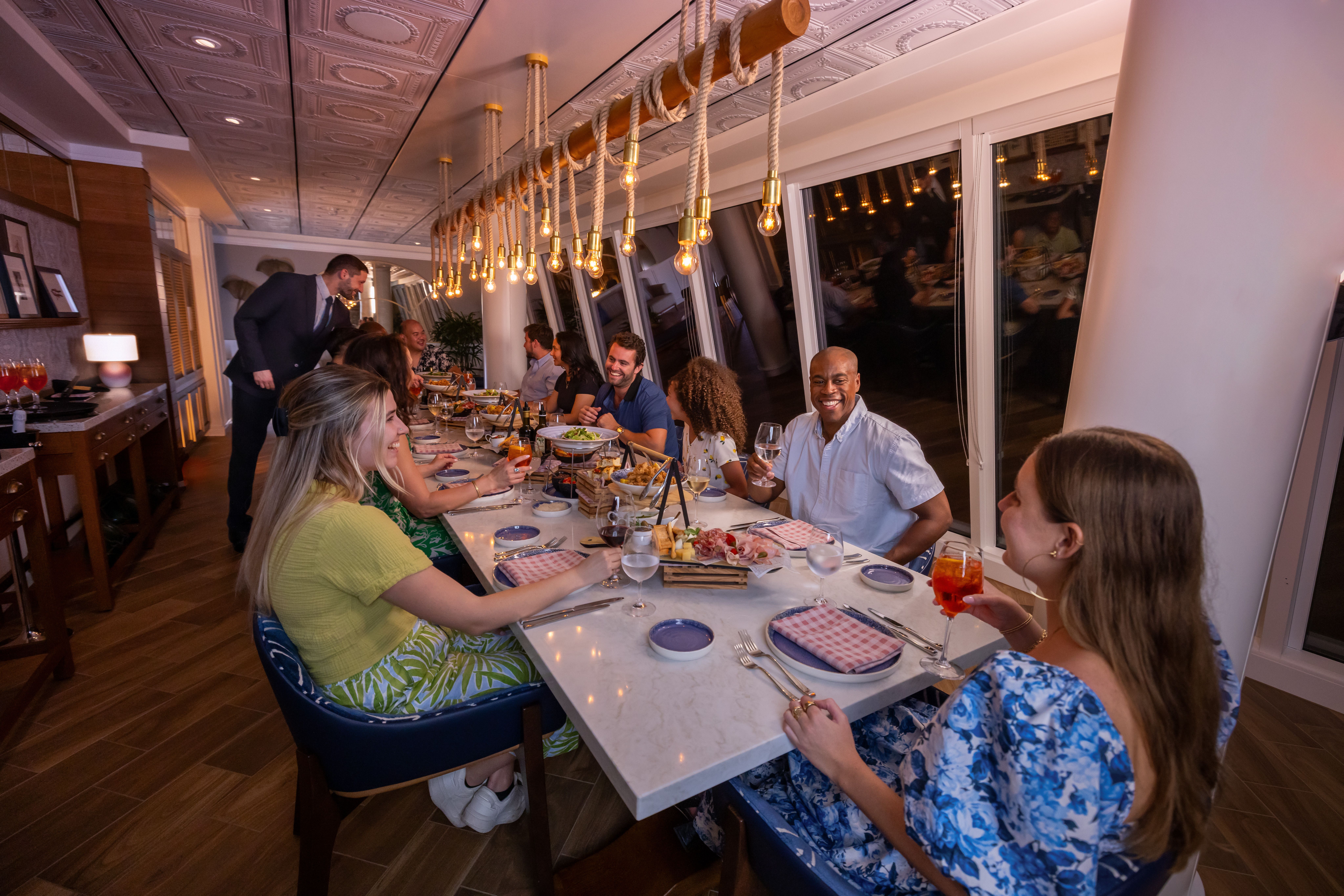 Group of friends enjoying dinner and drinks at a celebration table aboard Royal Caribbean, with gourmet dishes, drinks, and oceanview lighting.