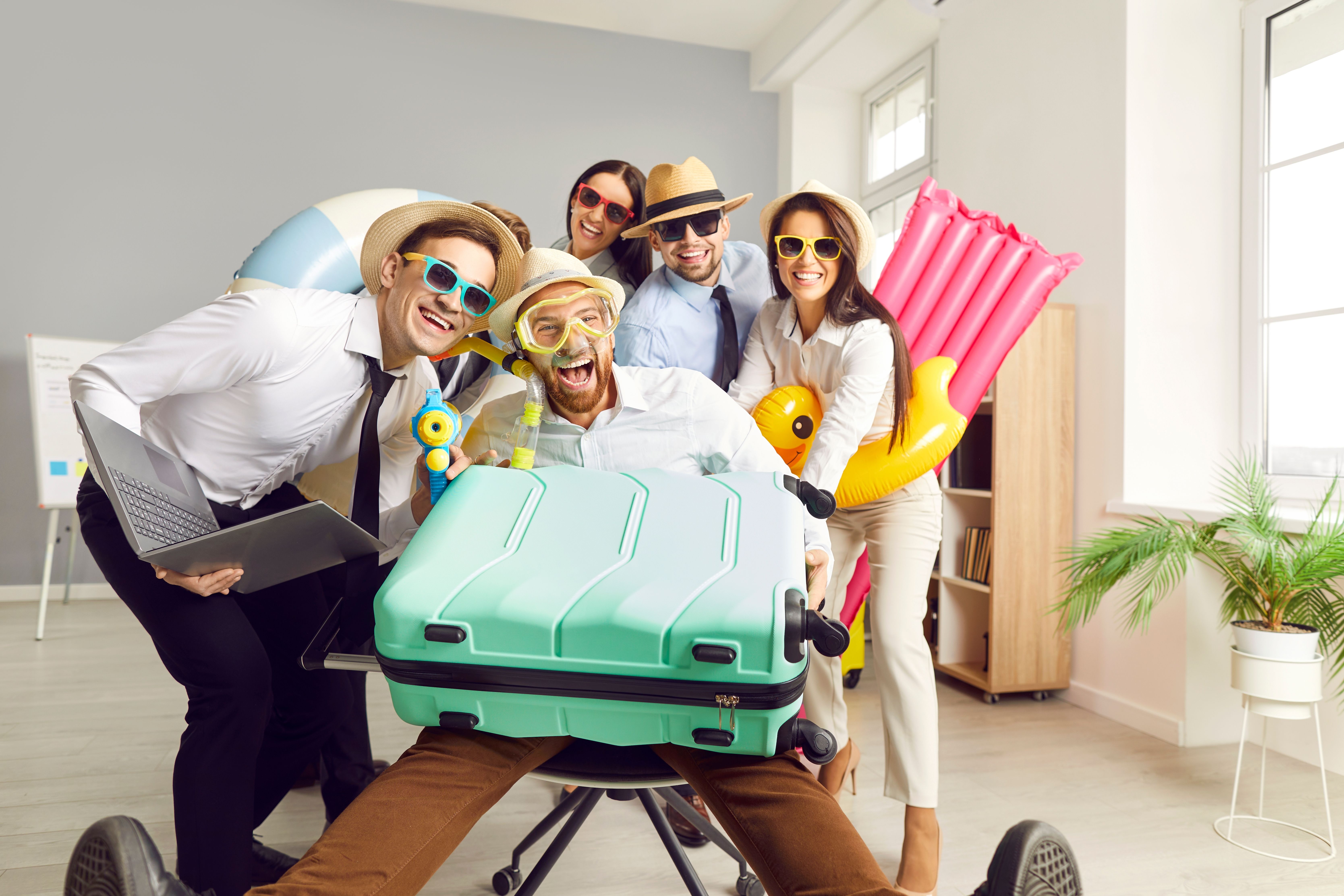 Coworkers dressed for fun planning a group vacation in the office with suitcase, laptop, and beach inflatables, symbolizing cruise getaway prep.