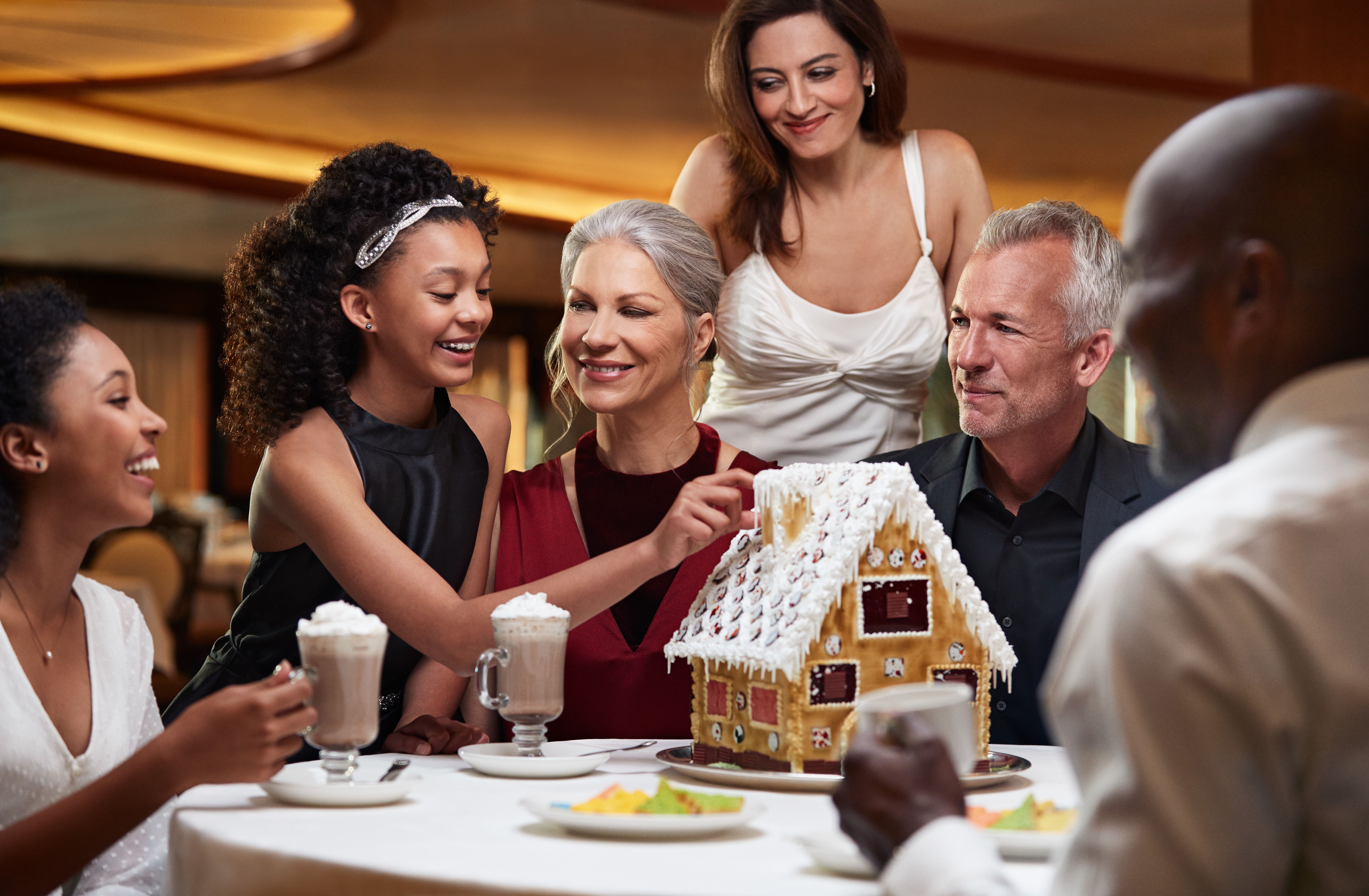 Multigenerational family building a gingerbread house and enjoying hot chocolate together during a festive holiday cruise celebration.