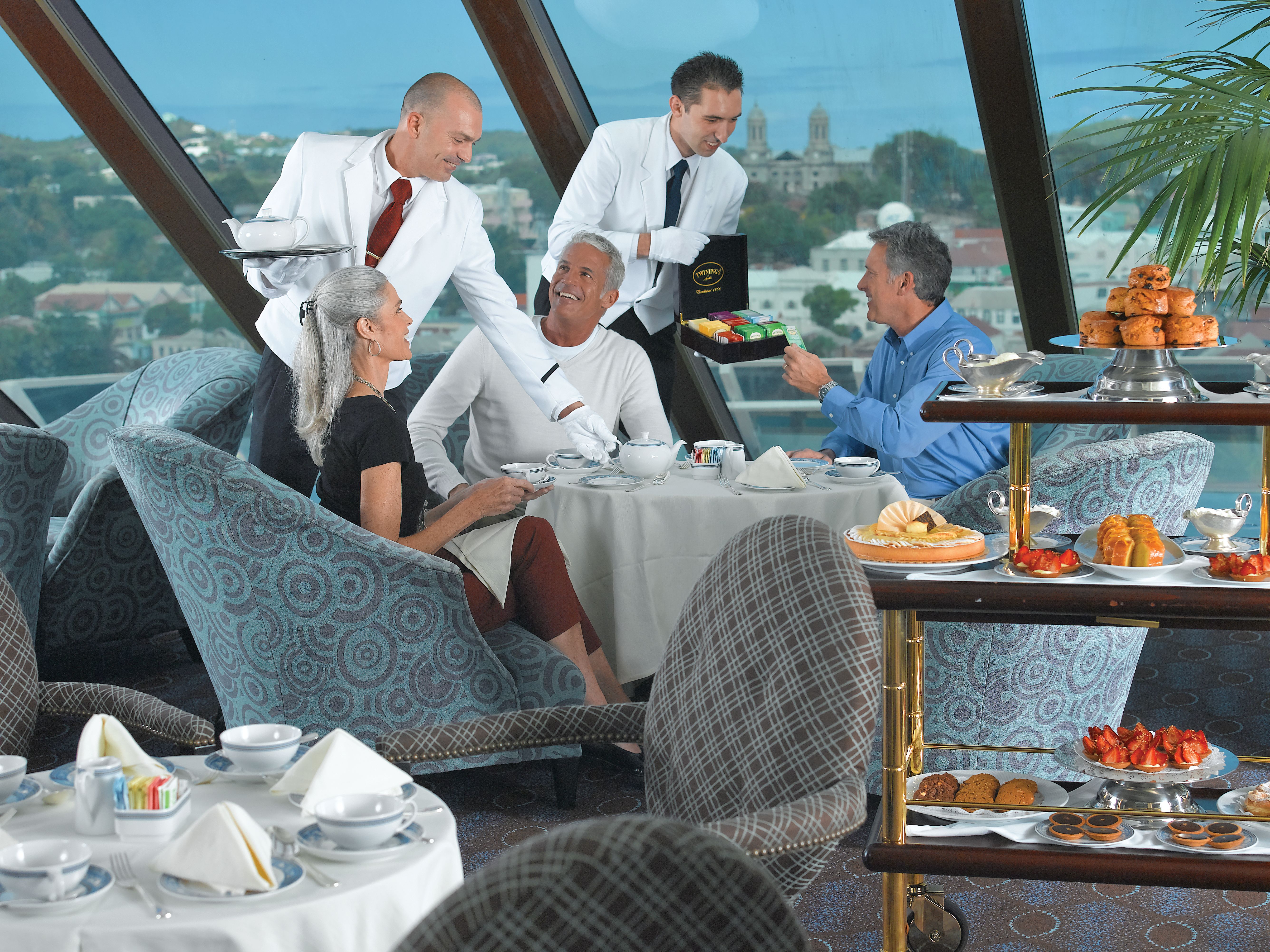 Guests enjoy afternoon tea in Horizons Lounge on Oceania Riviera with ocean views, elegant service, and a cart of pastries, scones, and premium tea selections.