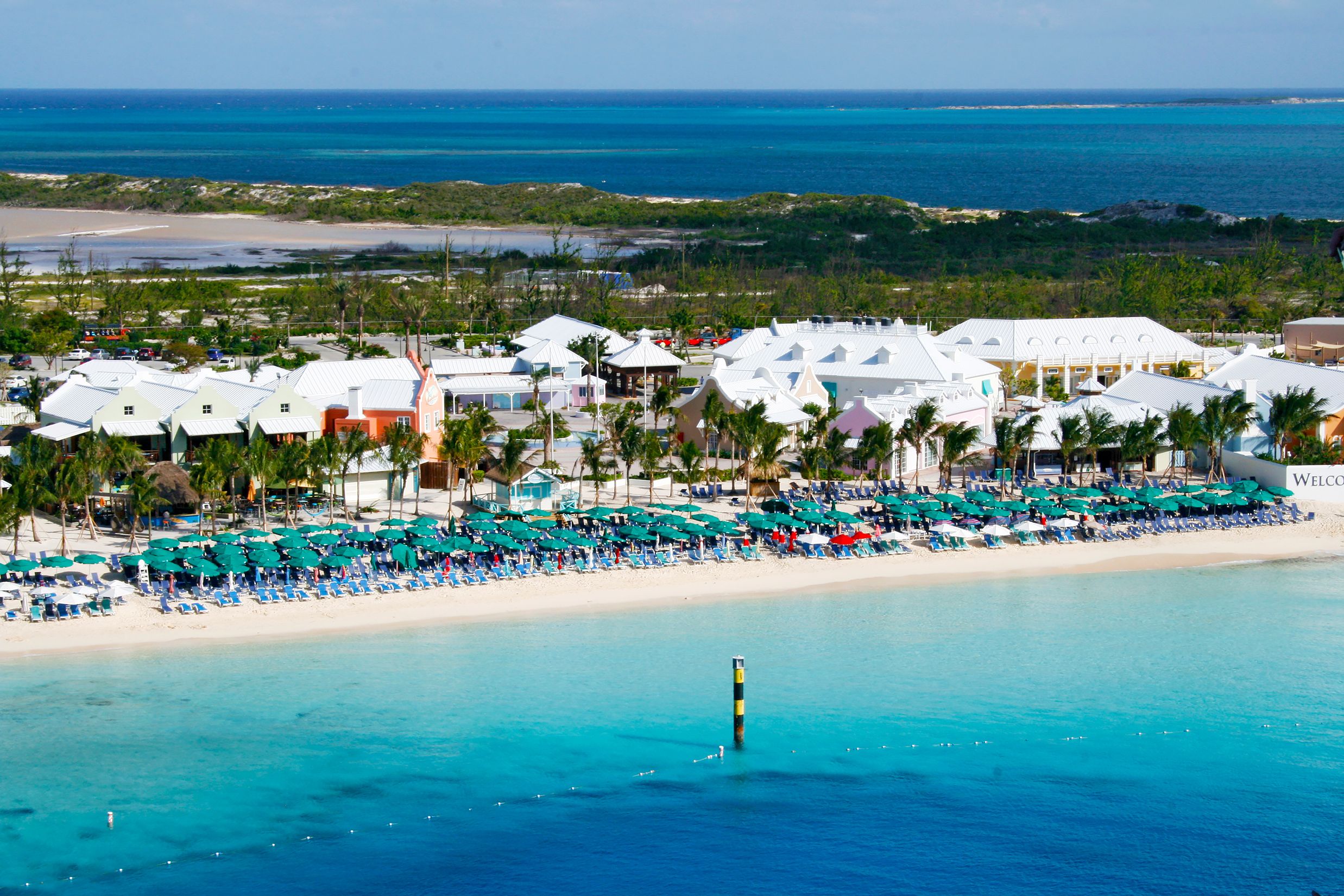 Aerial view of Margaritaville Beach Resort in Grand Turk, featuring turquoise waters, white-sand beach, rows of loungers, palm trees, and colorful coastal buildings.