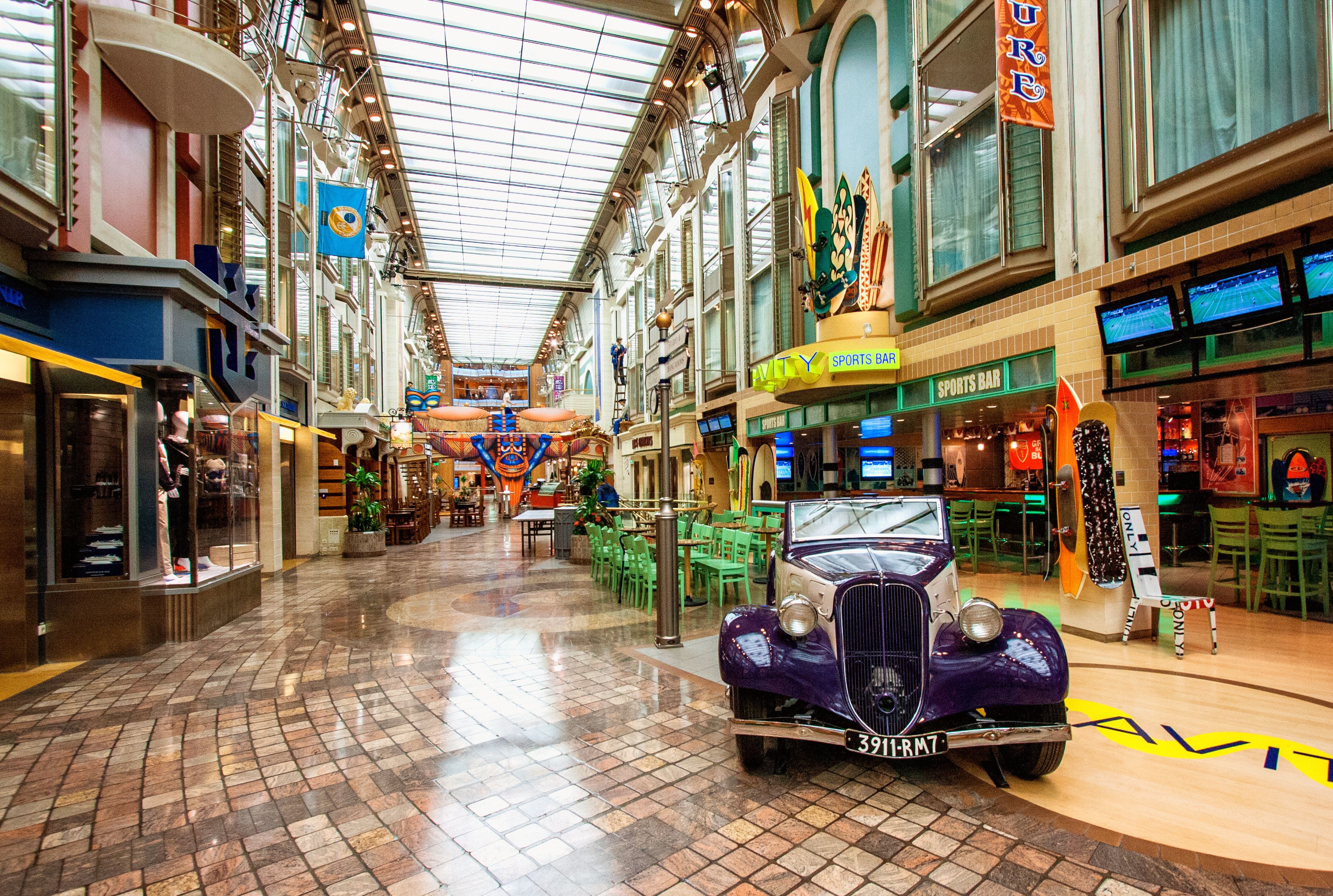 Royal Promenade aboard Adventure of the Seas featuring shops, sports bar, and vintage car display under a bright glass atrium with vibrant cruise decor.
