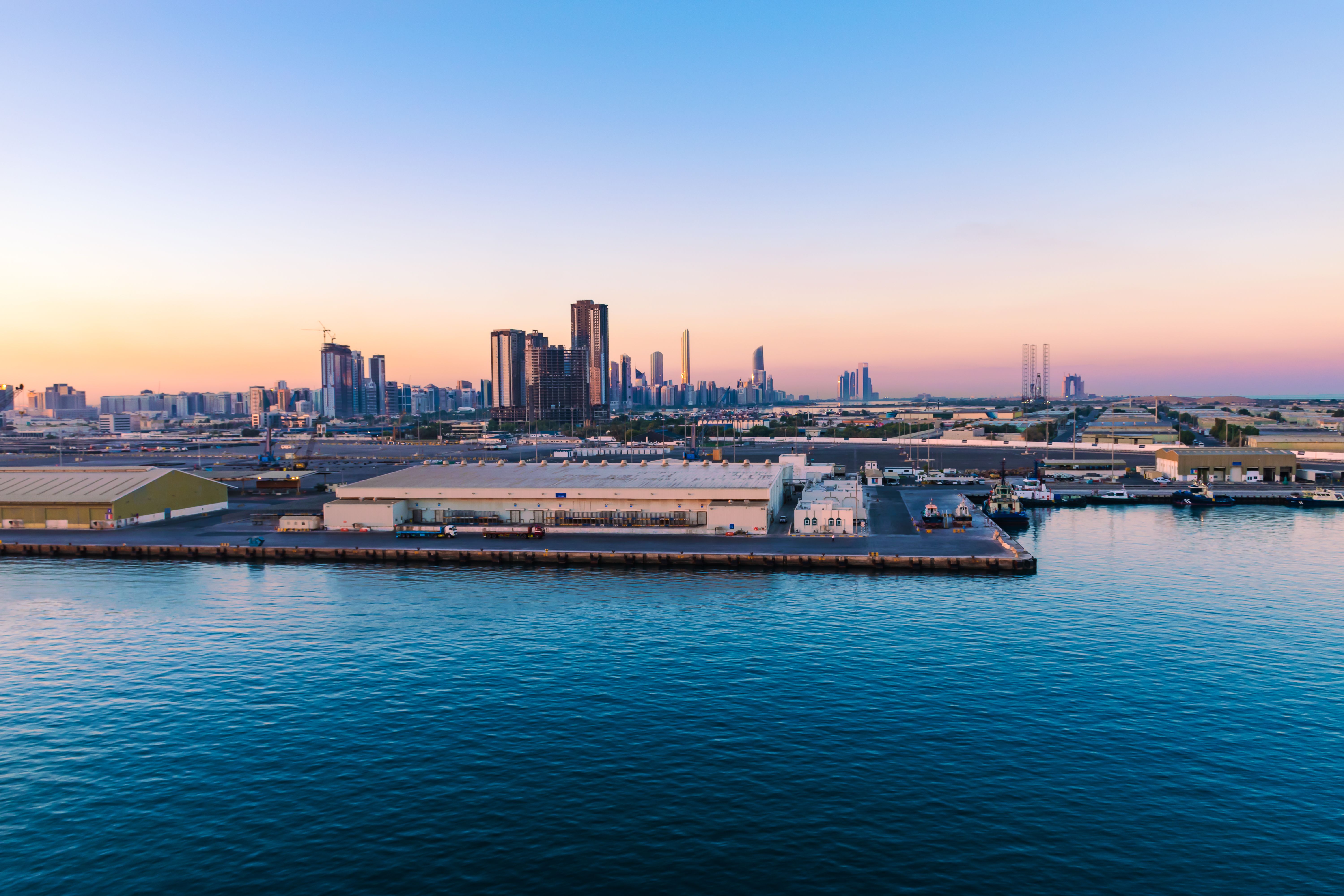 Abu Dhabi Cruise Terminal with city skyline at sunrise, showing port facilities and modern high-rises along the Arabian Gulf waterfront.