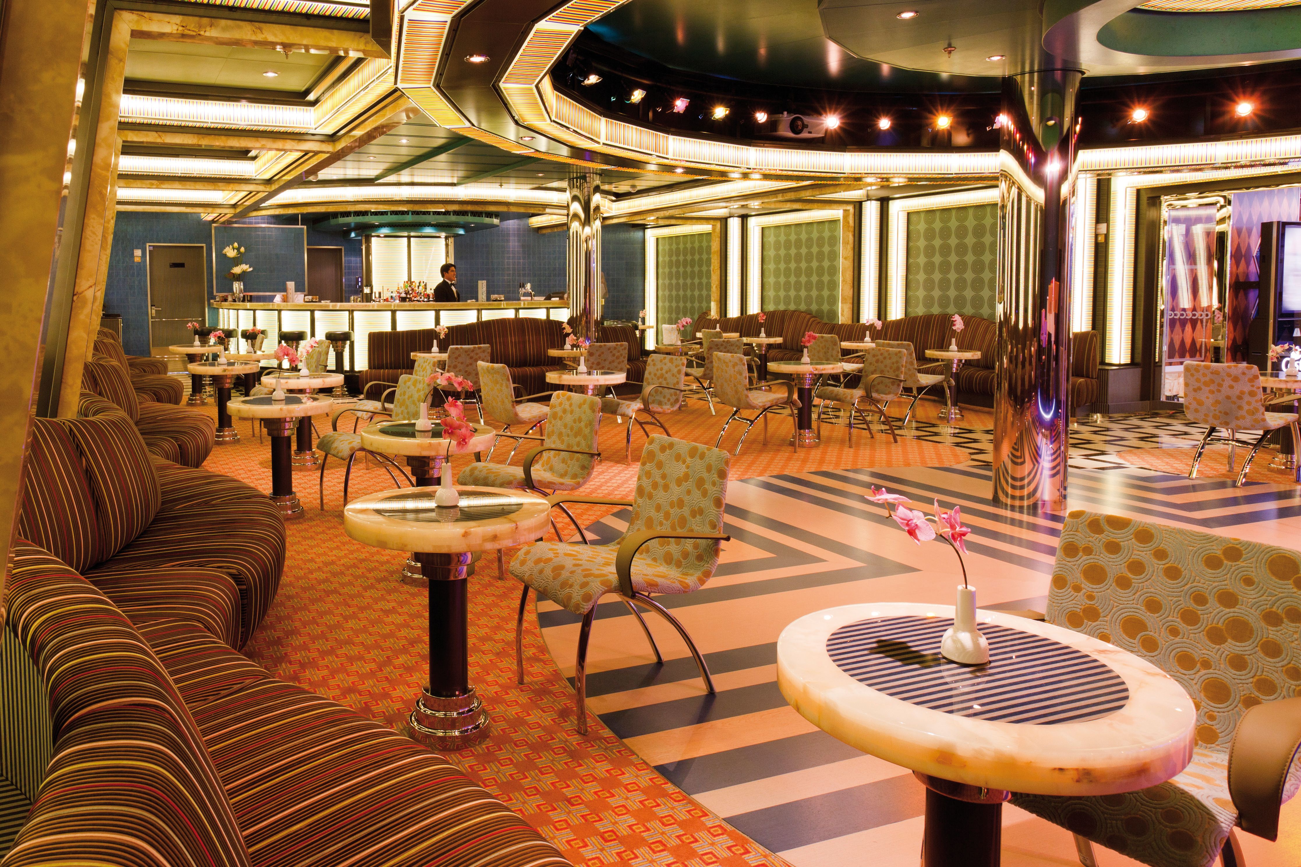 Costa Favolosa dance hall with art deco design, striped lounge seating, patterned chairs, marble cocktail tables, and a glowing bar in the background.