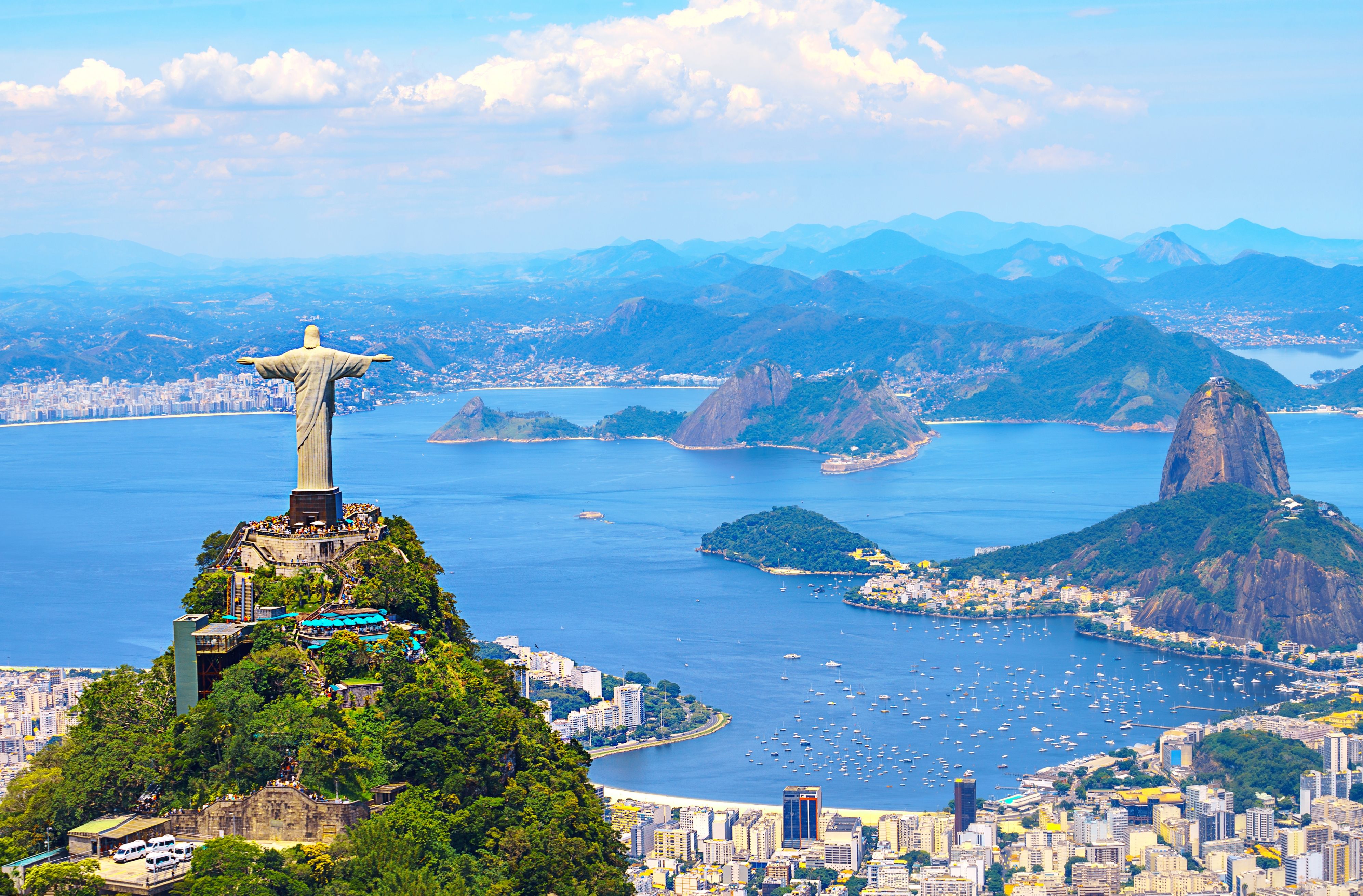 Christ the Redeemer overlooking Rio de Janeiro with Sugarloaf Mountain, Guanabara Bay, and city skyline under a clear blue sky.
