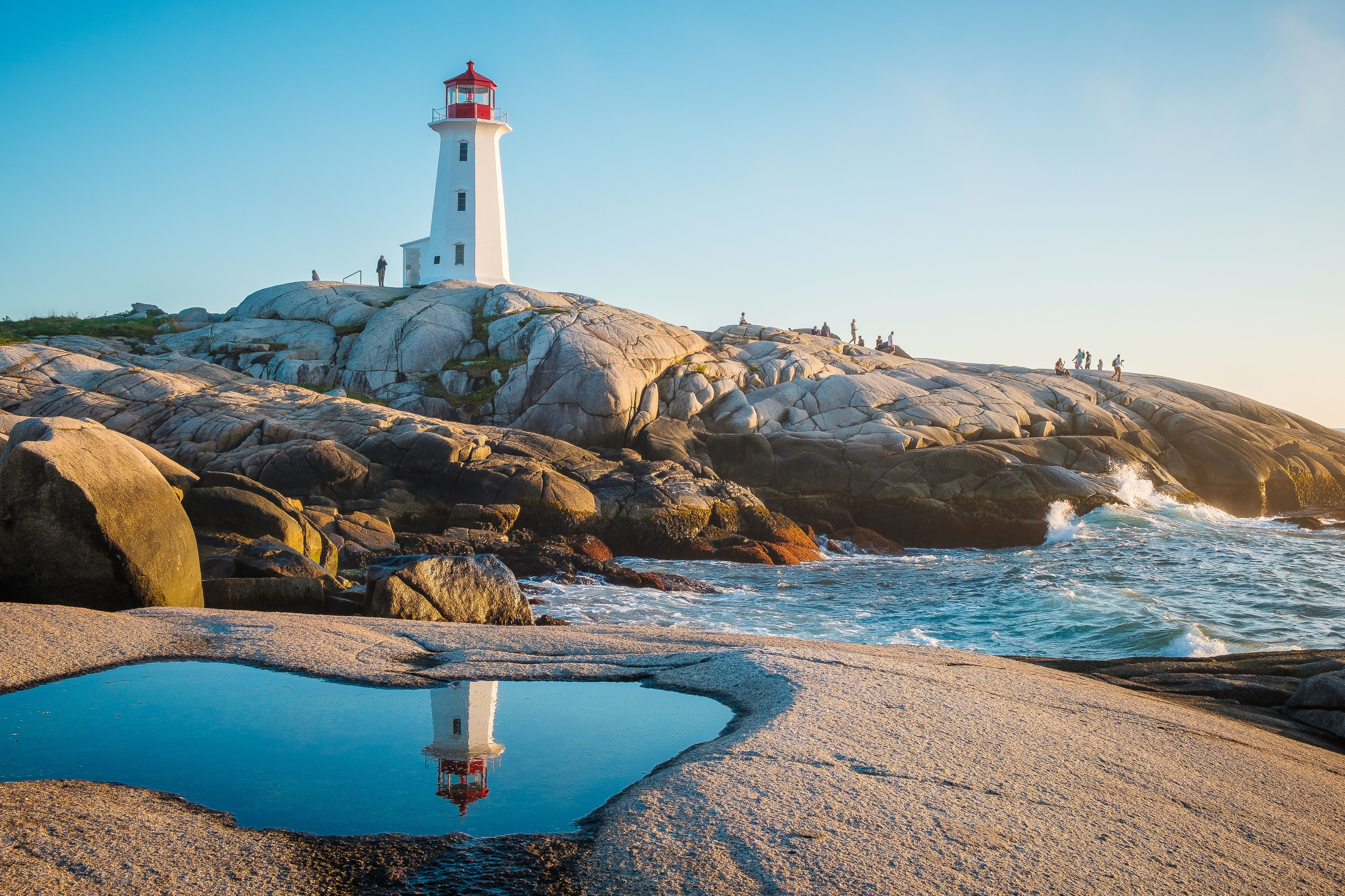 Peggy’s Cove Lighthouse in Nova Scotia atop smooth coastal rocks, reflected in tidal pool with ocean waves and golden hour sunlight.