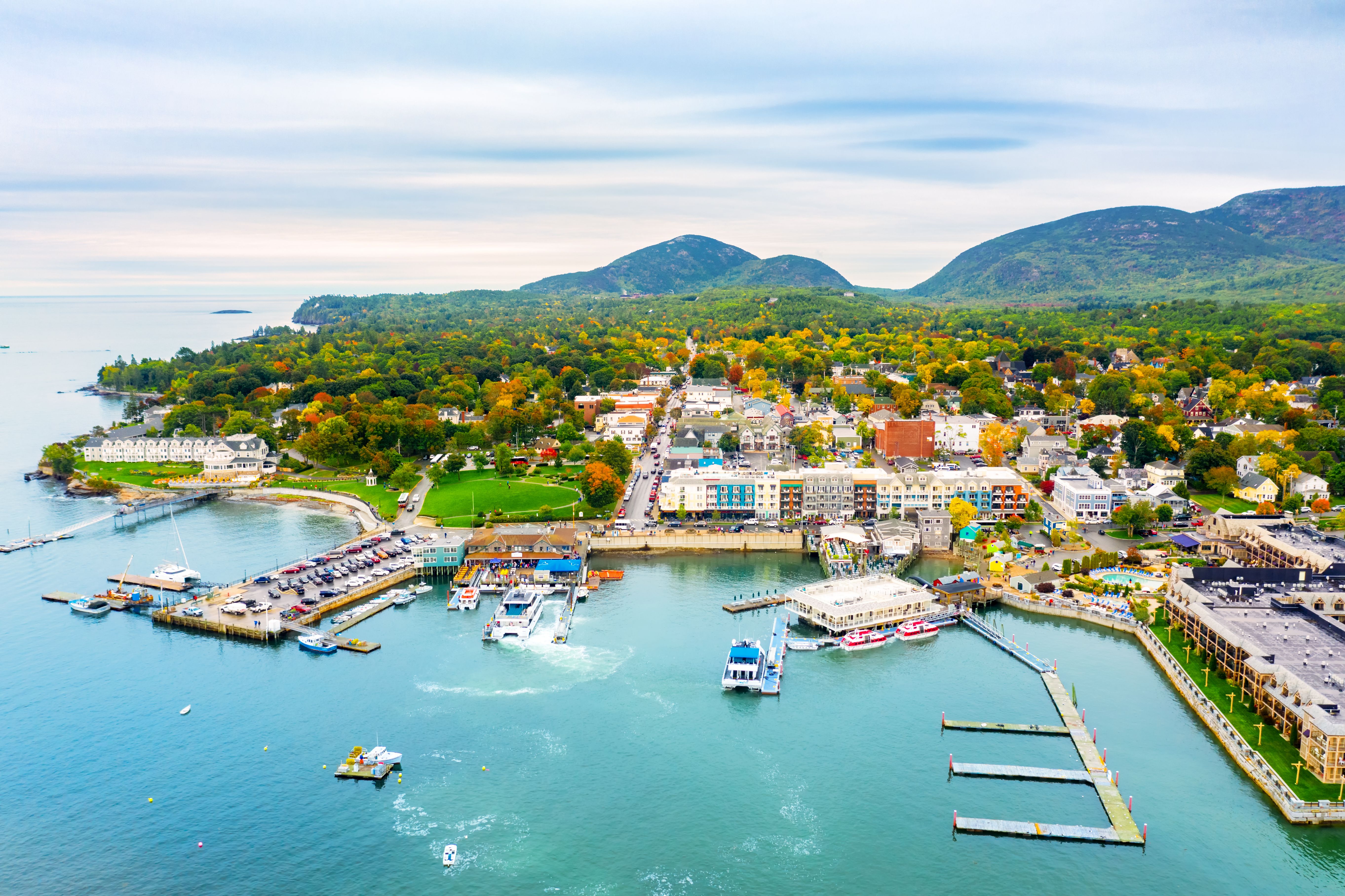Aerial view of Bar Harbor, Maine with colorful waterfront buildings, busy marina, forested hills, and fall foliage in the background.