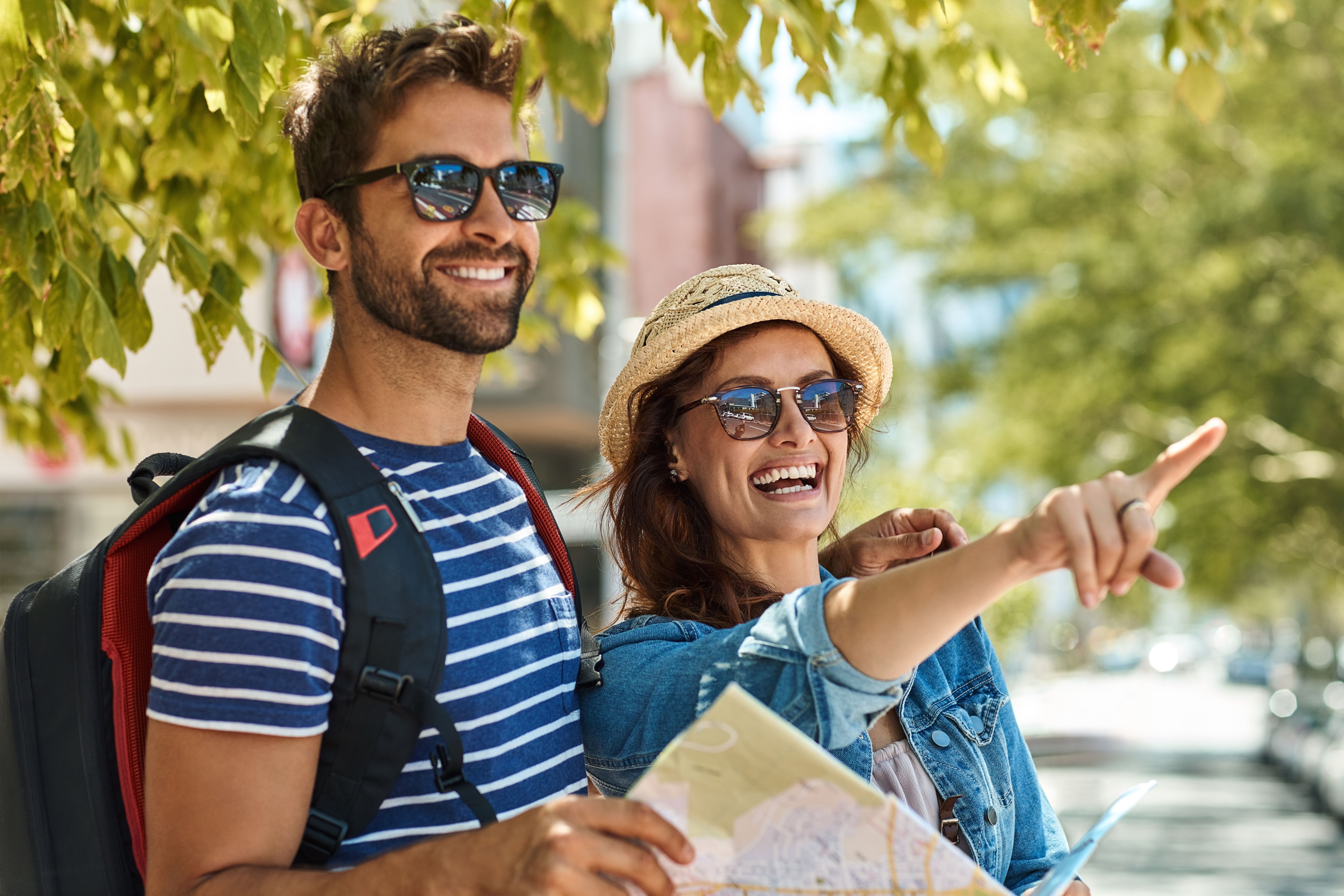 Smiling couple sightseeing in sunny weather, holding a map and pointing ahead, wearing sunglasses and casual travel attire under leafy green trees.