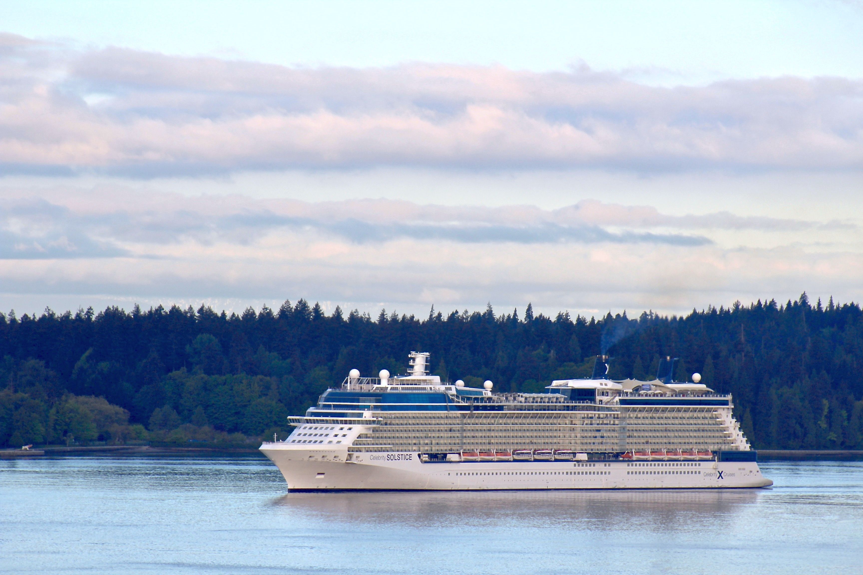 Celebrity Solstice cruise ship sailing near forested Canadian coastline under a cloudy sky, reflecting on calm coastal waters.