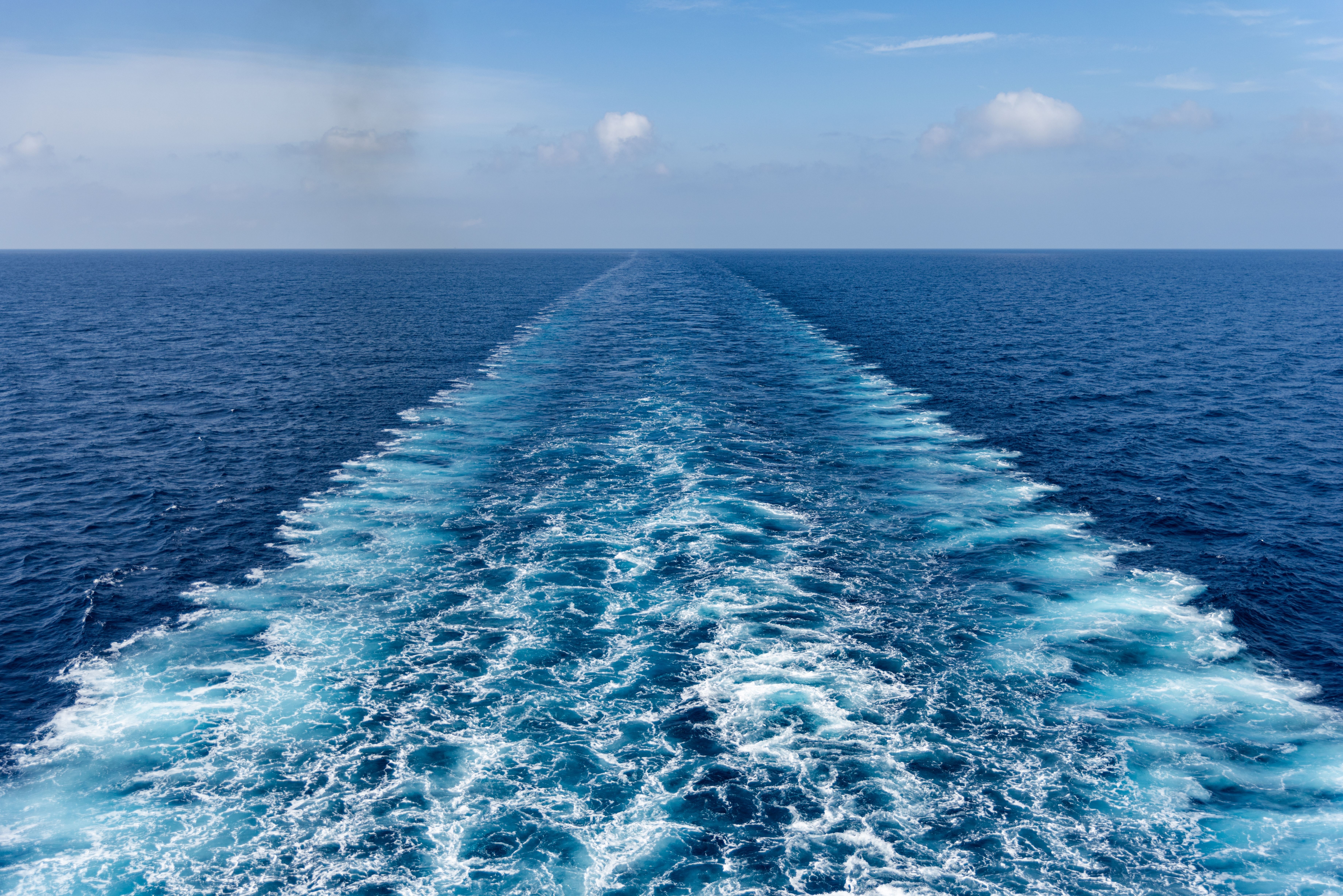 Foamy stern wake behind a cruise ship in calm blue ocean under a partly cloudy sky.