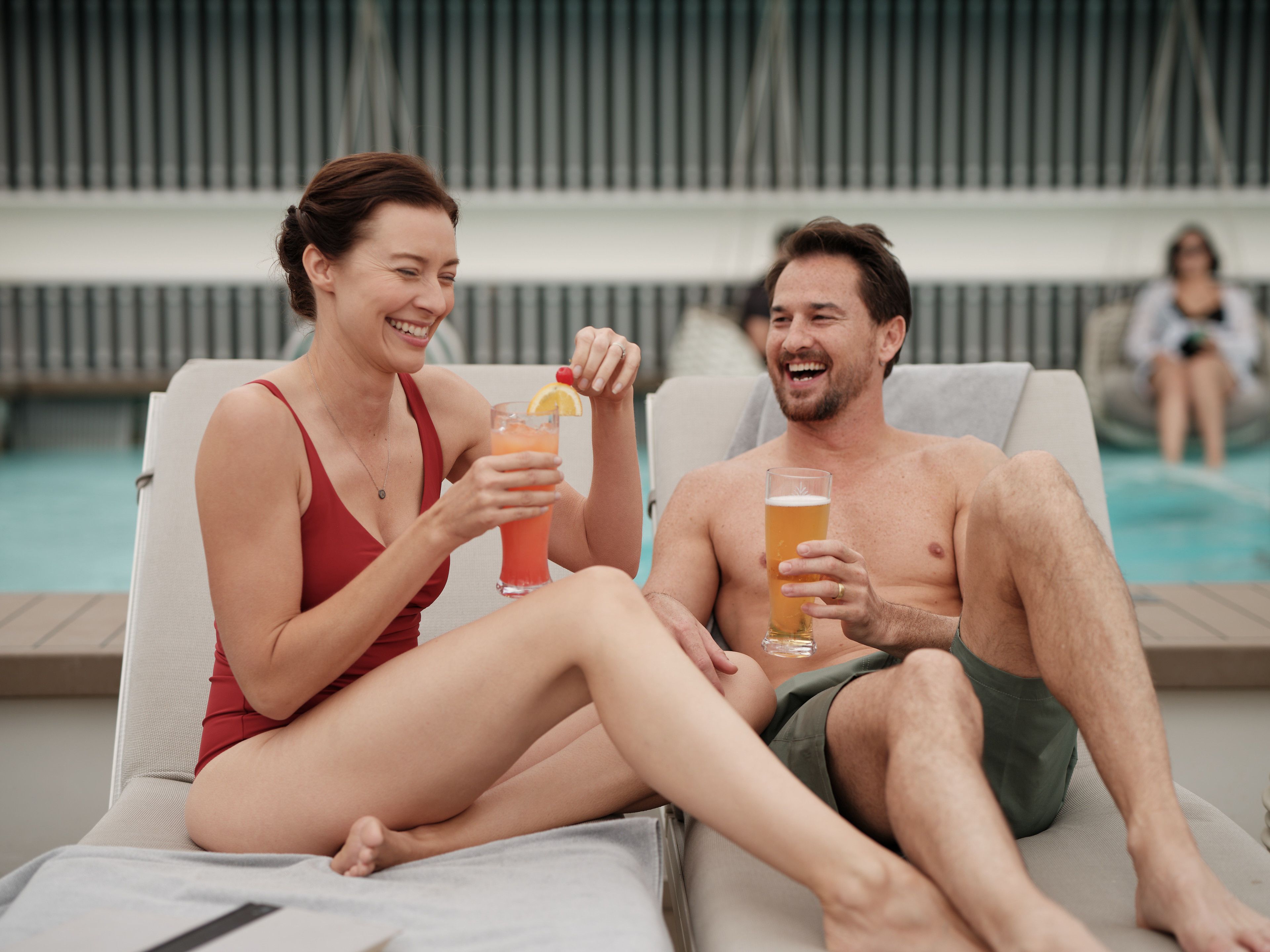Couple lounging by the cruise ship pool, smiling and enjoying cocktails in swimwear on a cloudy day.