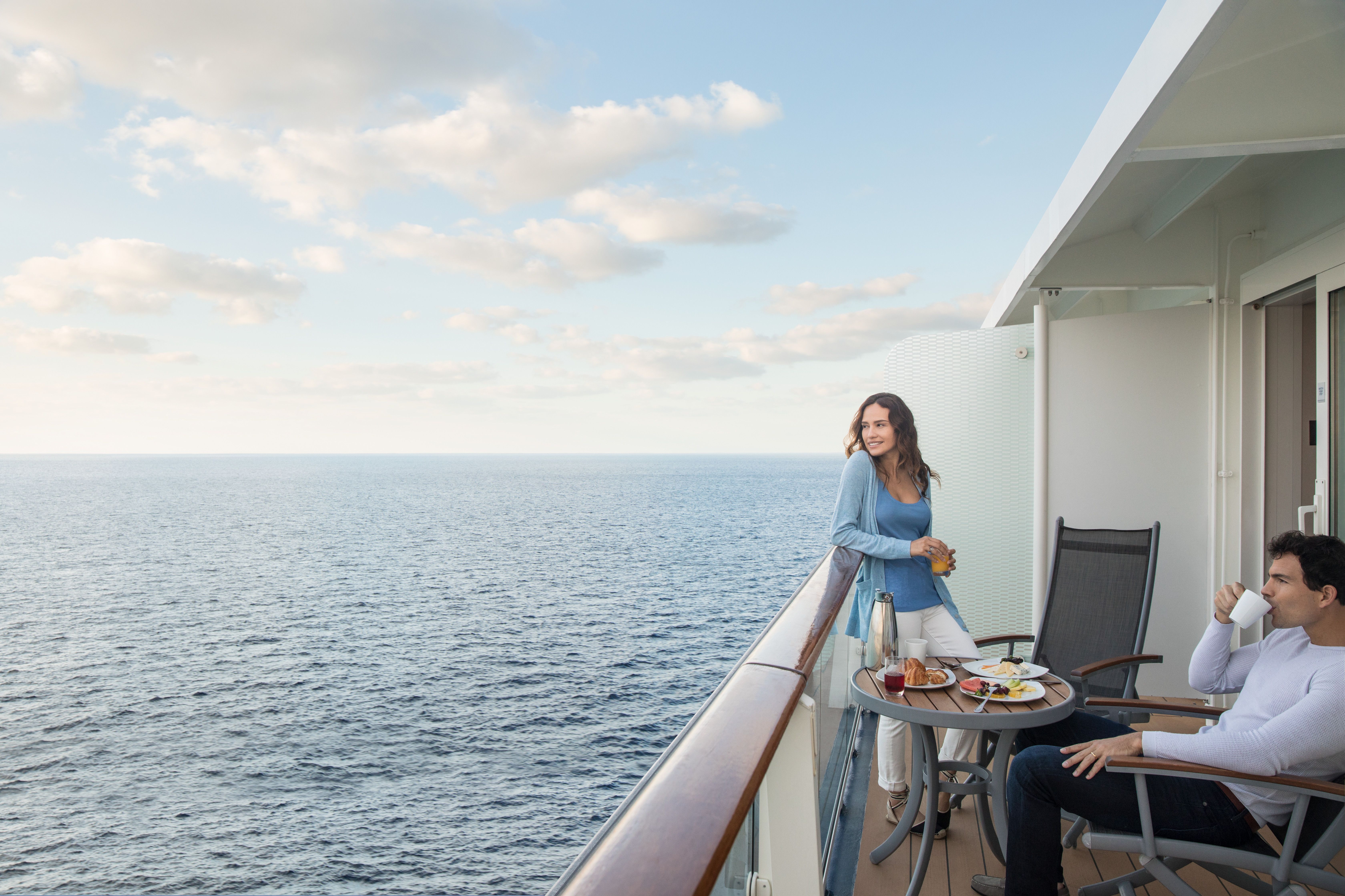 Couple enjoying breakfast and ocean views on a private cruise ship balcony, relaxing under a blue sky with scattered clouds.