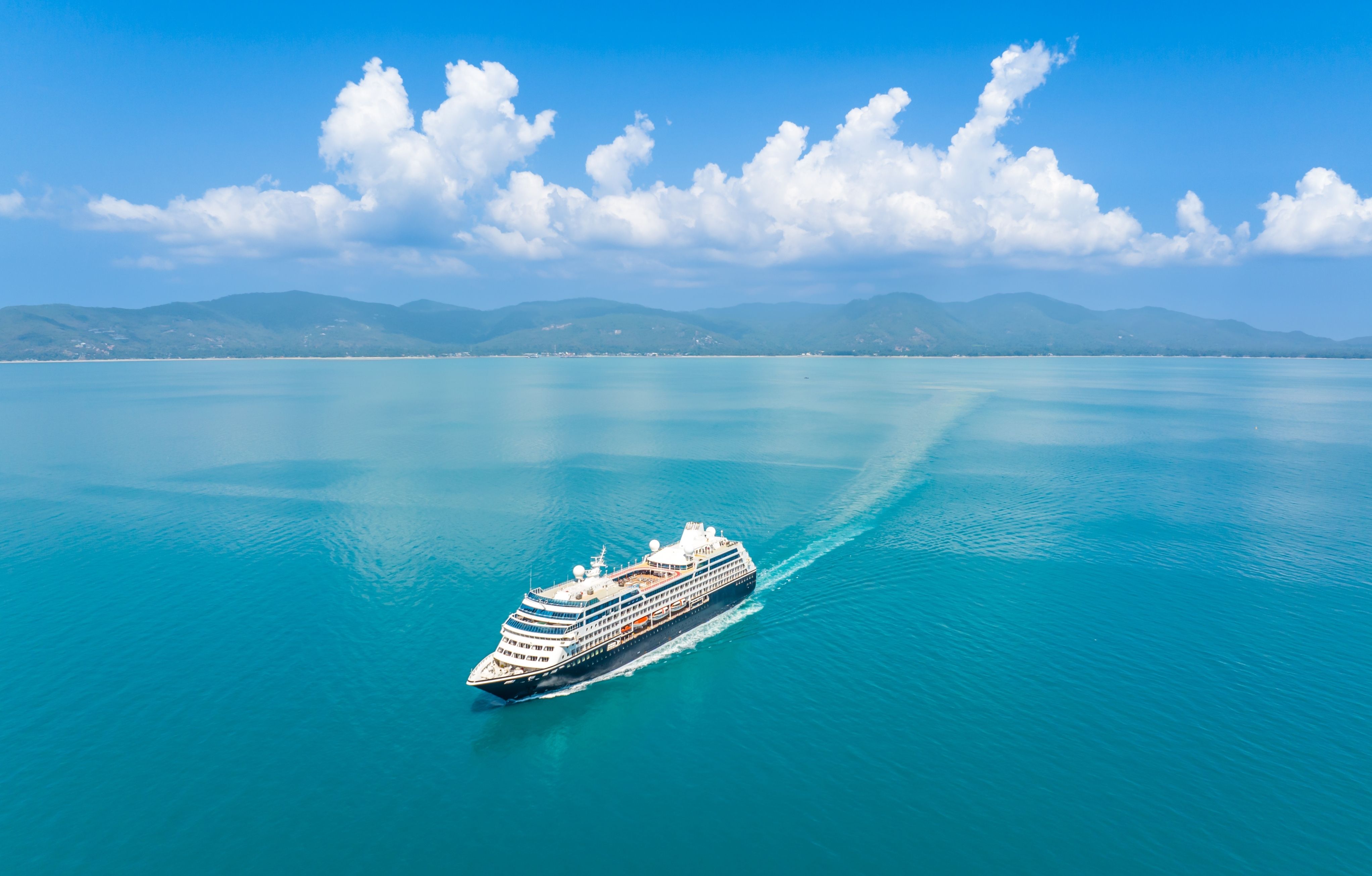 Luxury cruise ship sailing through calm turquoise waters with mountainous coastline and blue sky in the background.