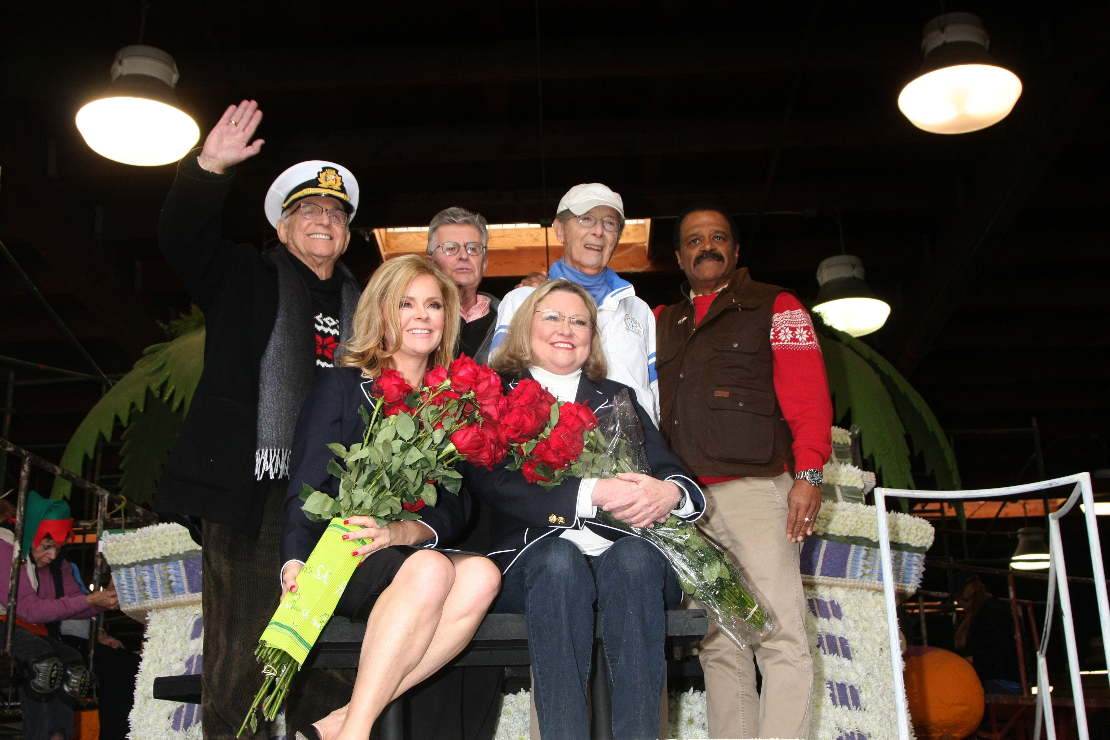 The Love Boat cast posing with roses aboard a decorated float, celebrating at an indoor event with festive lighting and floral arrangements.