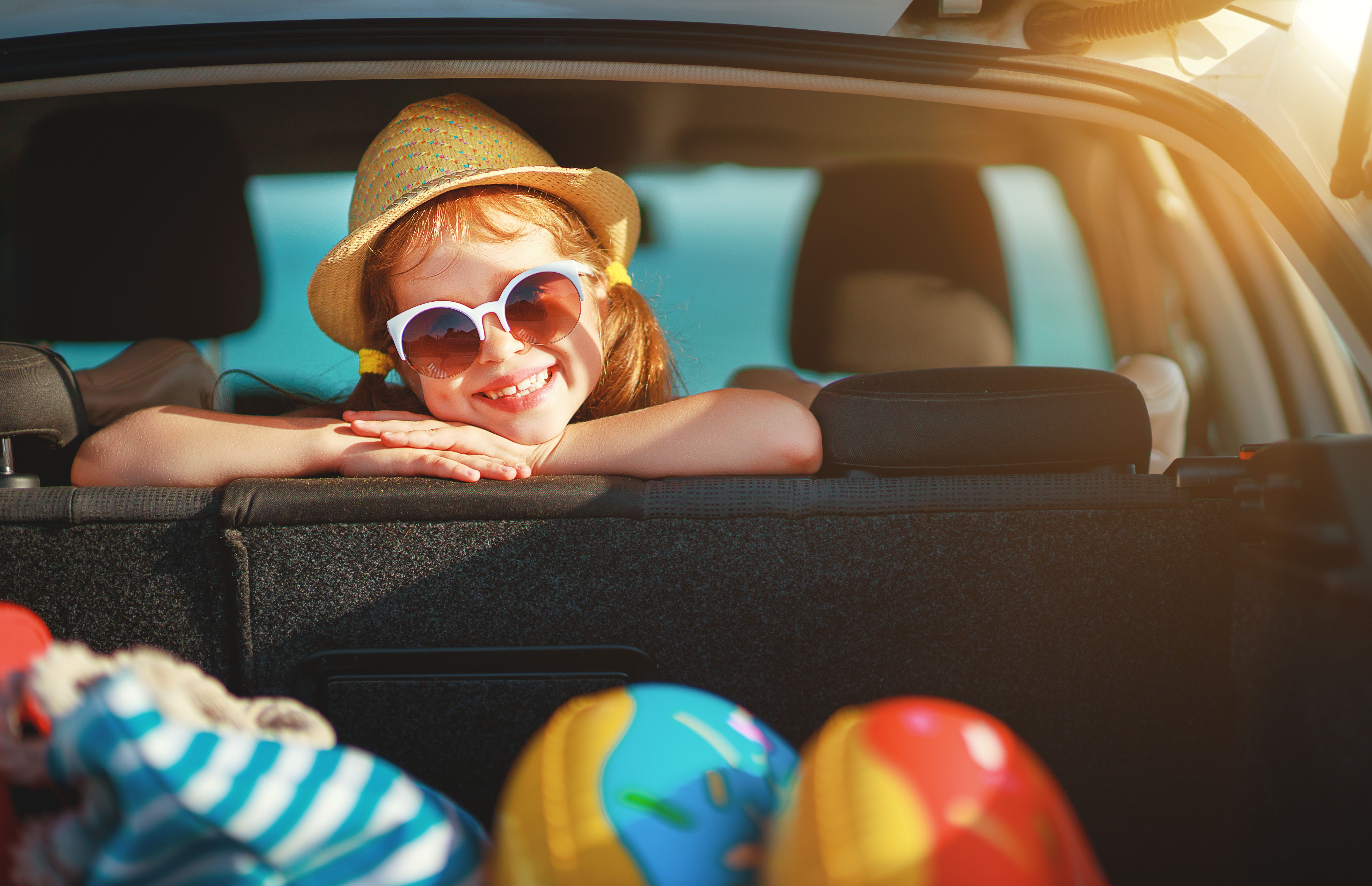 Smiling child in sunglasses and hat leans from car trunk packed for summer vacation, perfect for promoting family-friendly drive-to cruise ports.