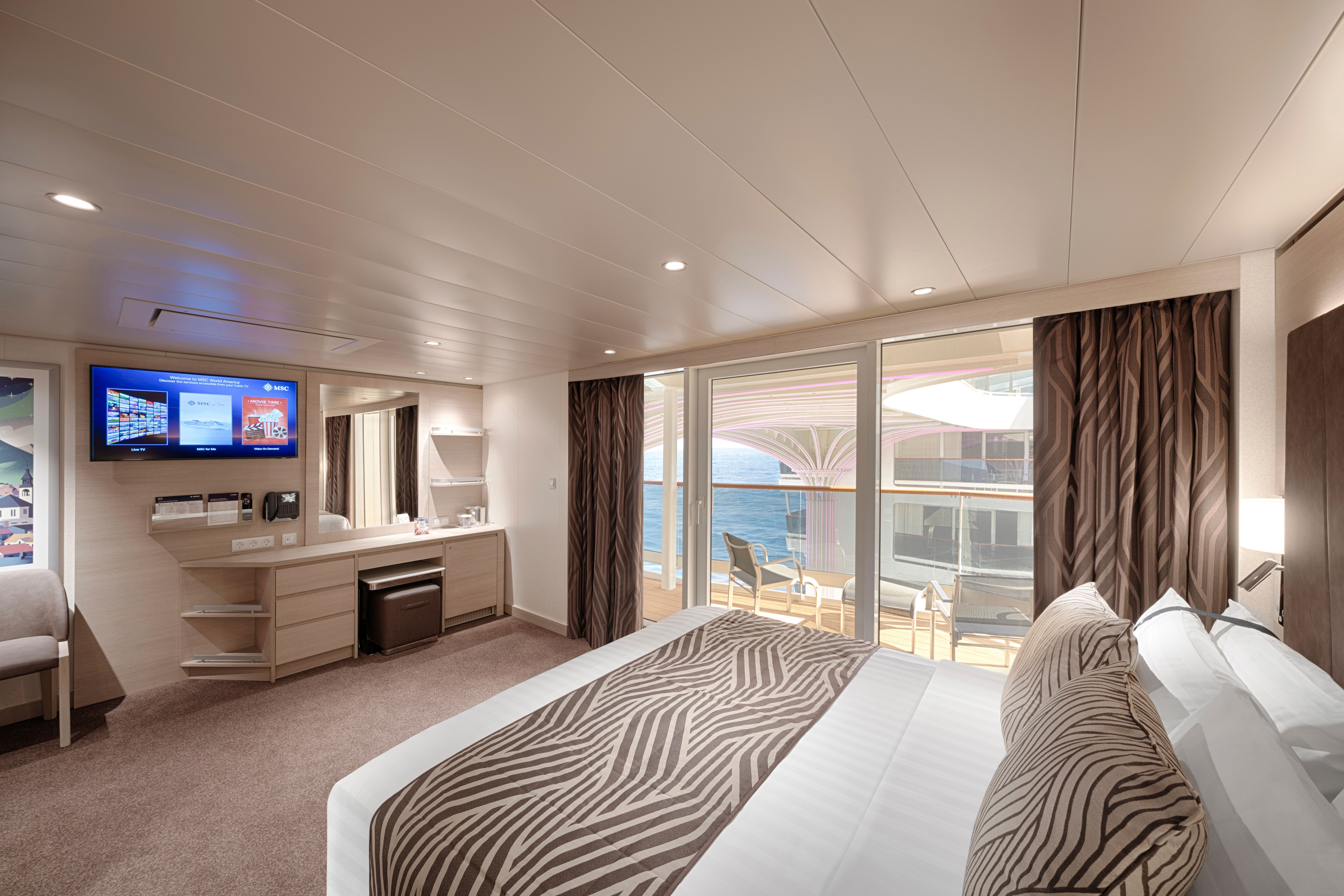 Elegant MSC World America Junior Suite Aurea with large bed, flat-screen TV, and direct access to a stylish balcony overlooking sea and promenade.