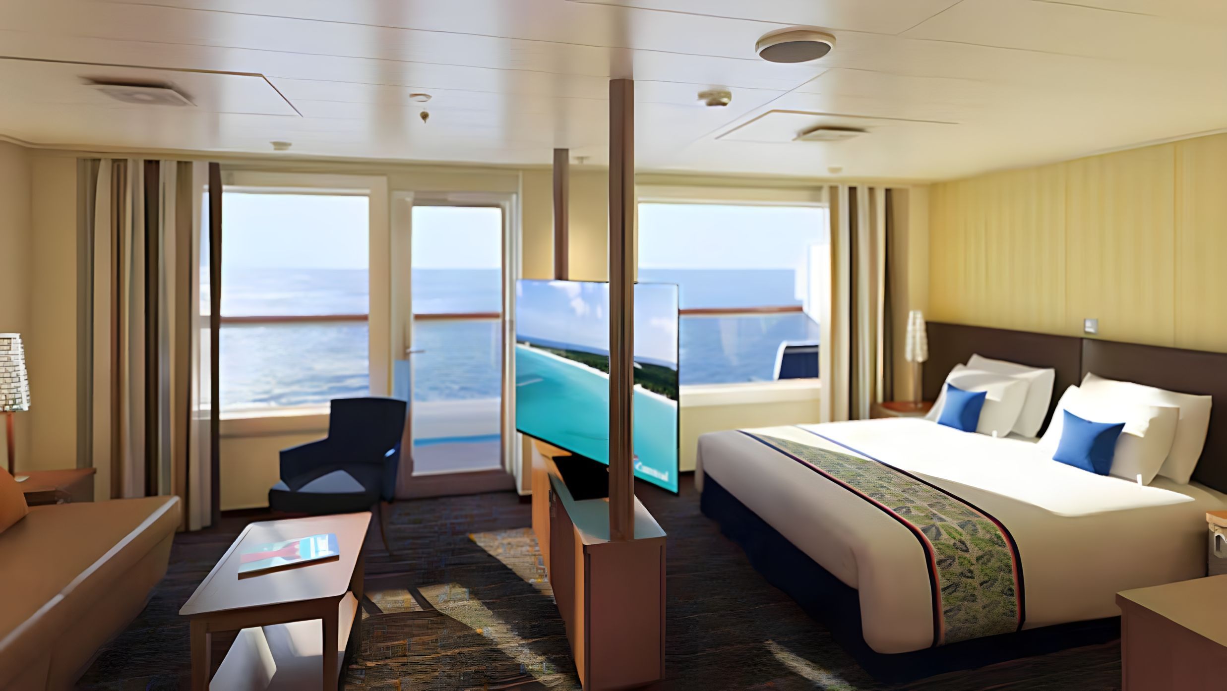 Carnival Grand Suite with king bed, separate living area, sofa, dining table, floor-to-ceiling windows, private balcony, and panoramic ocean views.