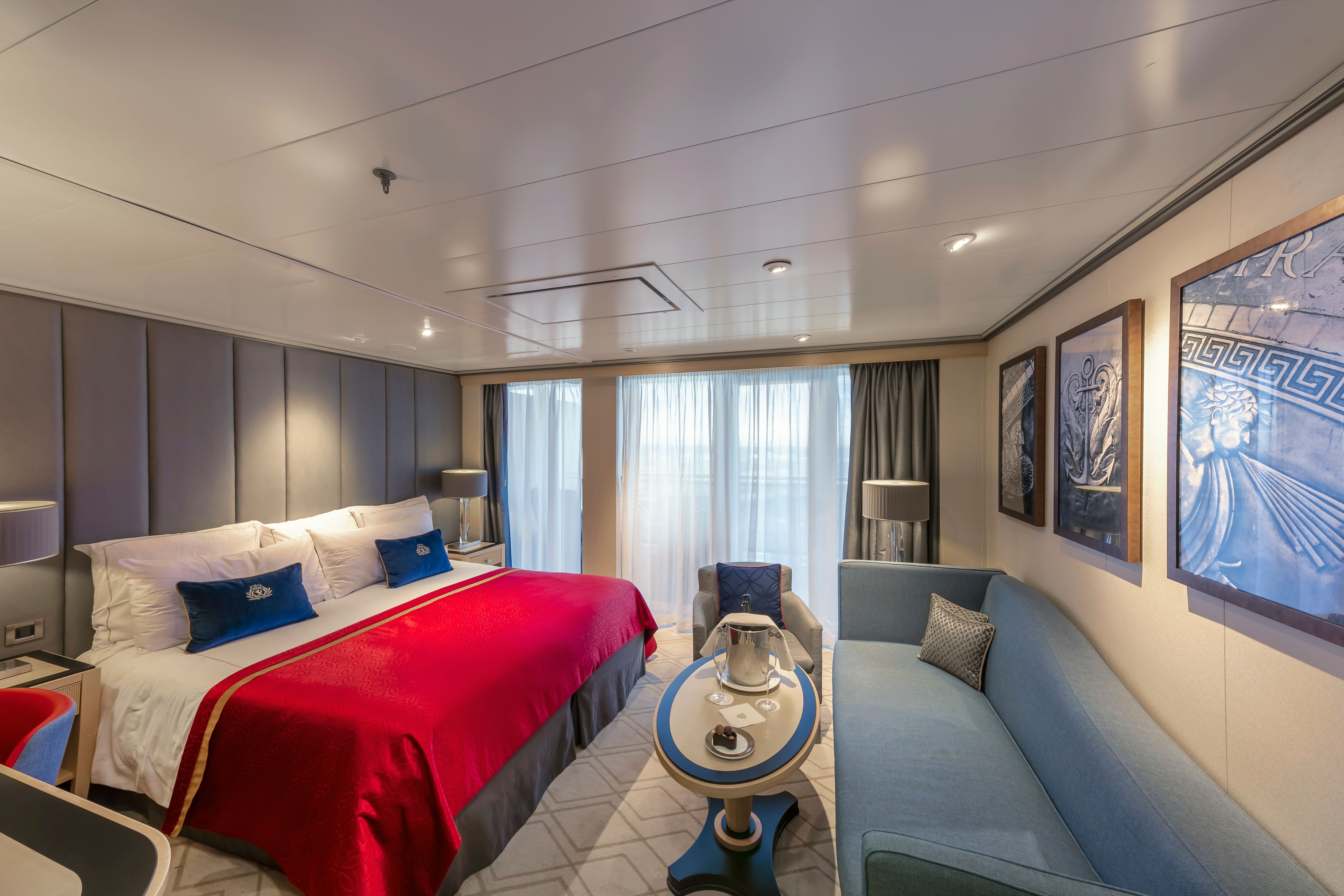 Queen Mary 2 Princess Suite bedroom with king-size bed, red accent throw, blue seating area, and elegant ocean liner décor.