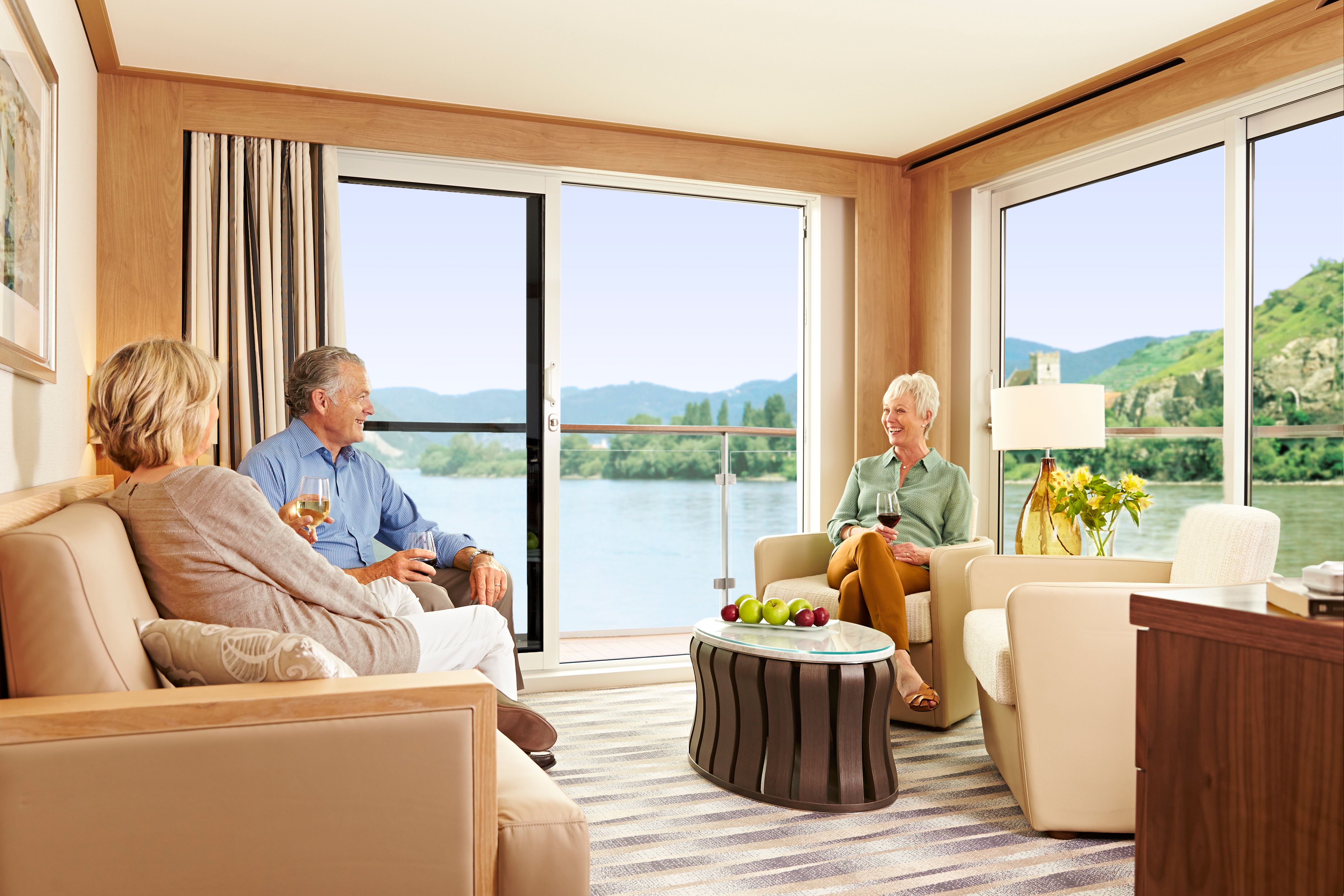 Viking River Cruises Explorer Suite living area with floor-to-ceiling windows and balcony views of a scenic European river valley, luxury river ship suite lounge with guests enjoying wine.
