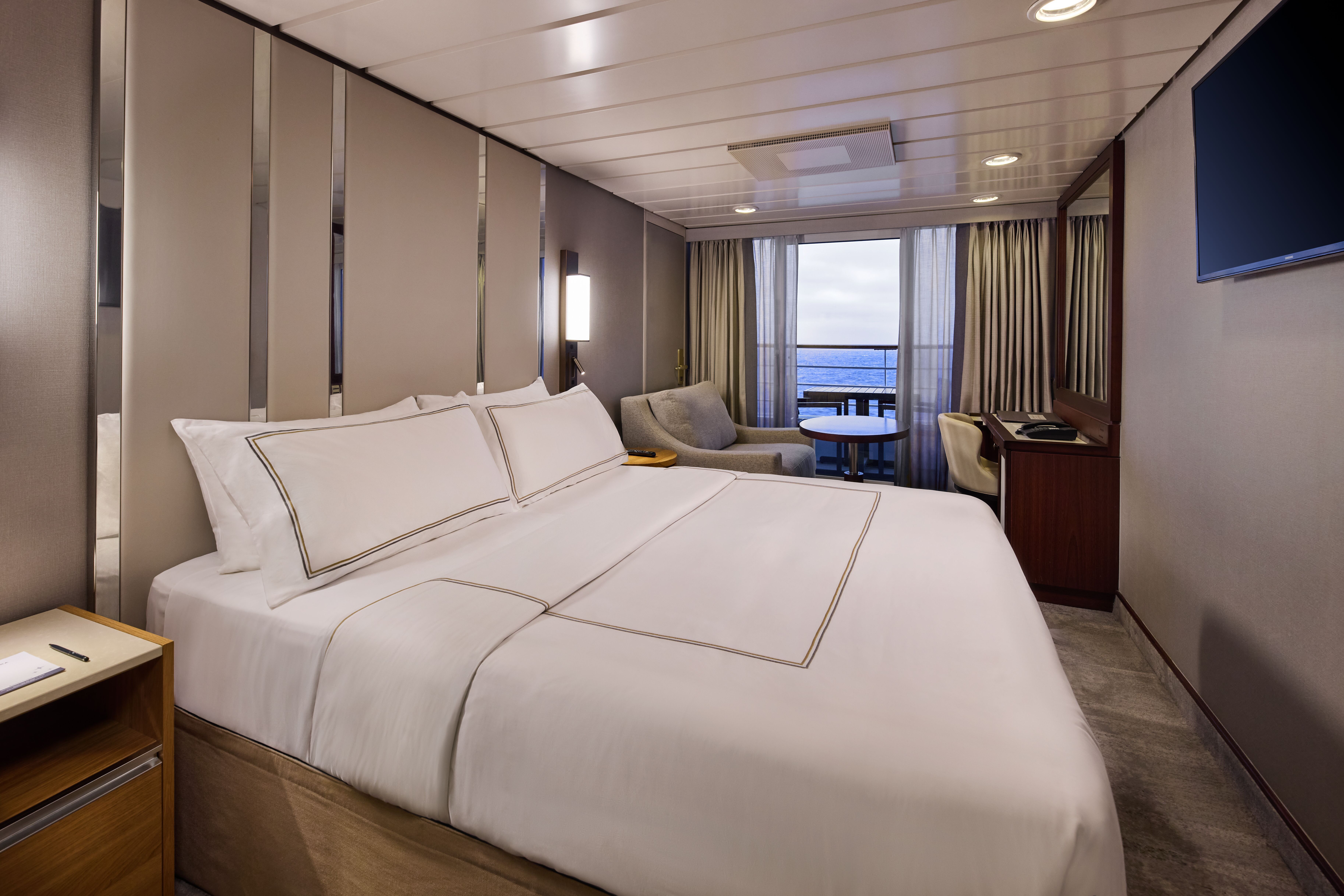 Azamara Pursuit Veranda Stateroom with plush queen bed, sitting area with armchair and sofa, vanity desk, wall-mounted TV, and private balcony with ocean views.