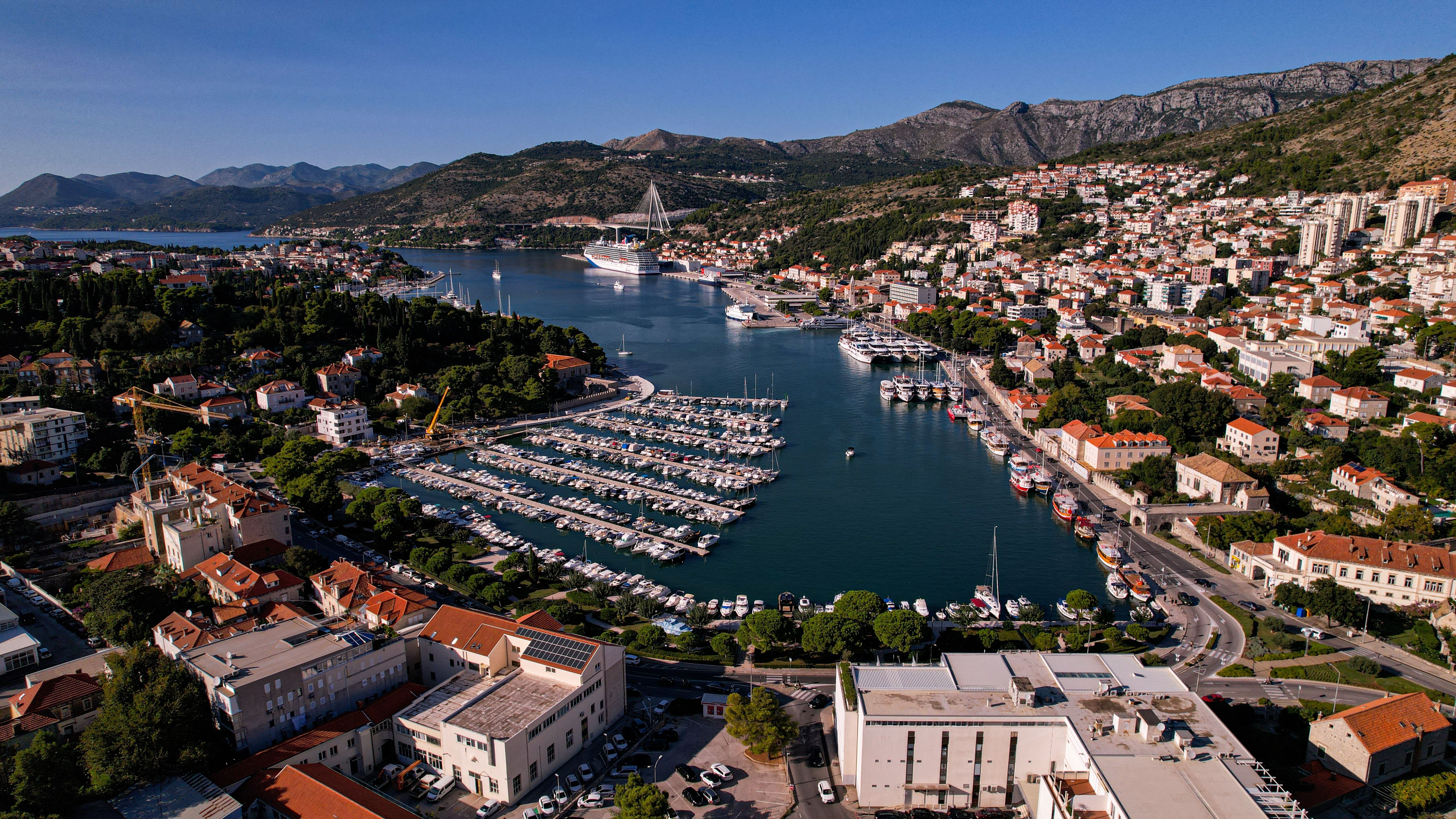 Dubrovnik cruise port aerial view with docked cruise ship, marina filled with yachts, Adriatic coastline, red-roof Old Town, and surrounding Dalmatian mountains in Croatia