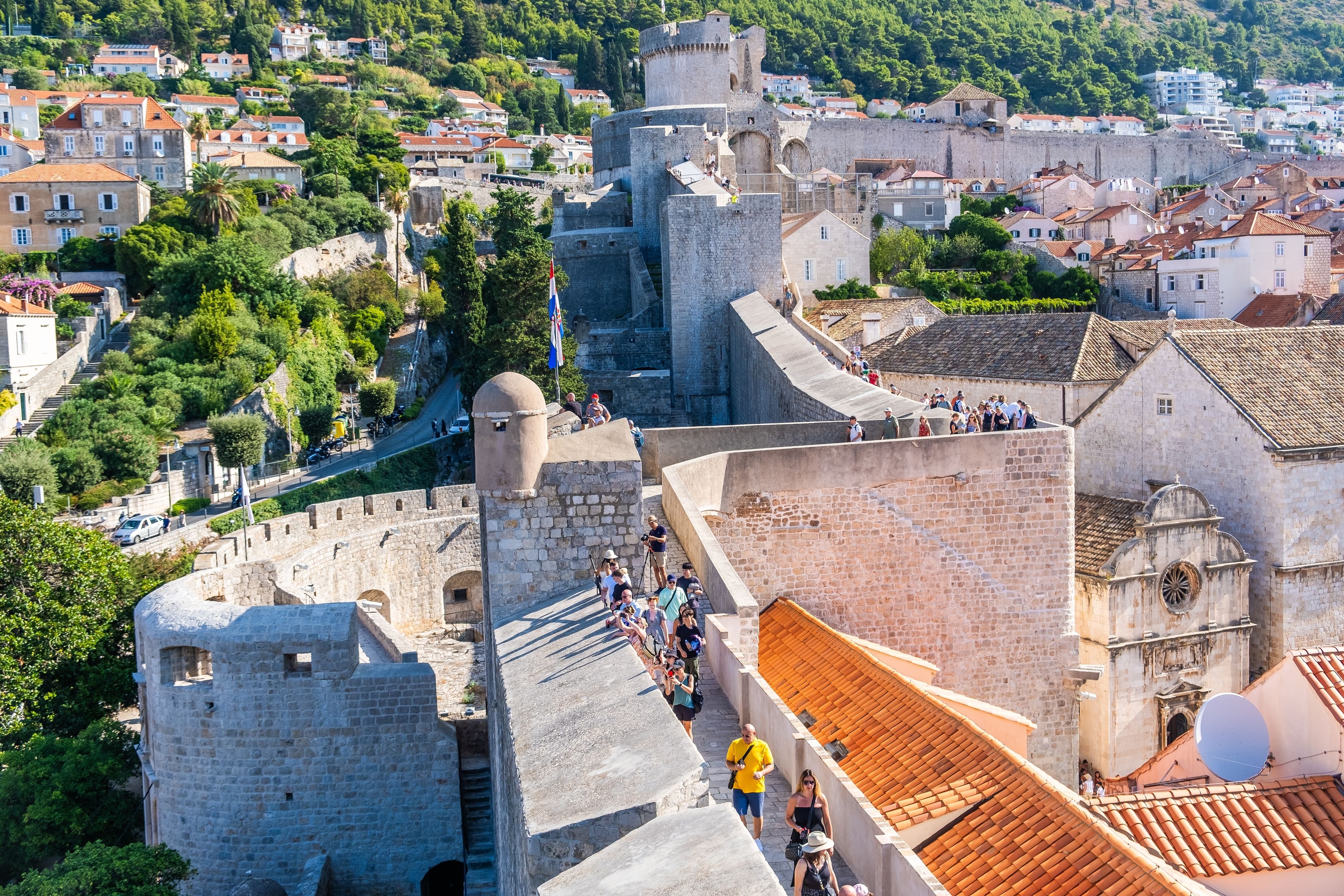 Dubrovnik Old Town city walls with walking tourists, medieval stone fortifications, terracotta rooftops and lush hillside backdrop, Croatia cruise shore excursion highlight