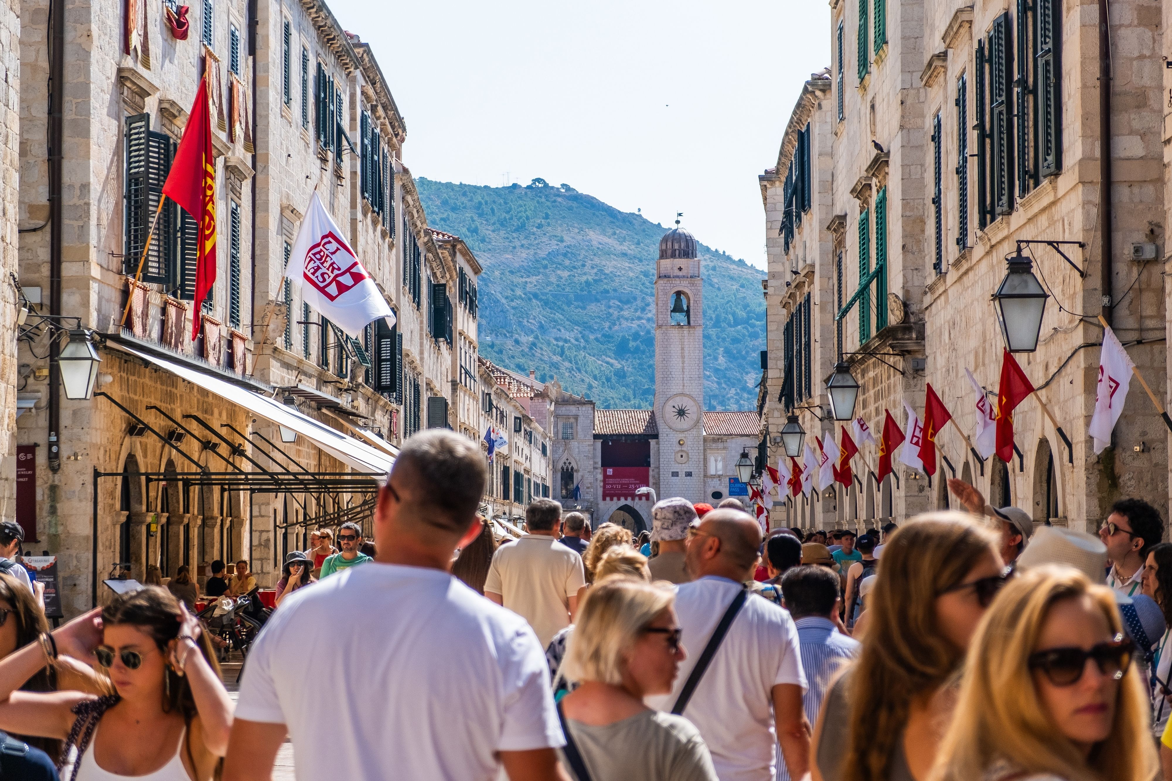 Dubrovnik Stradun street with crowds of tourists, historic limestone buildings, clock tower and flags, vibrant Old Town promenade, Croatia cruise port walking tour scene