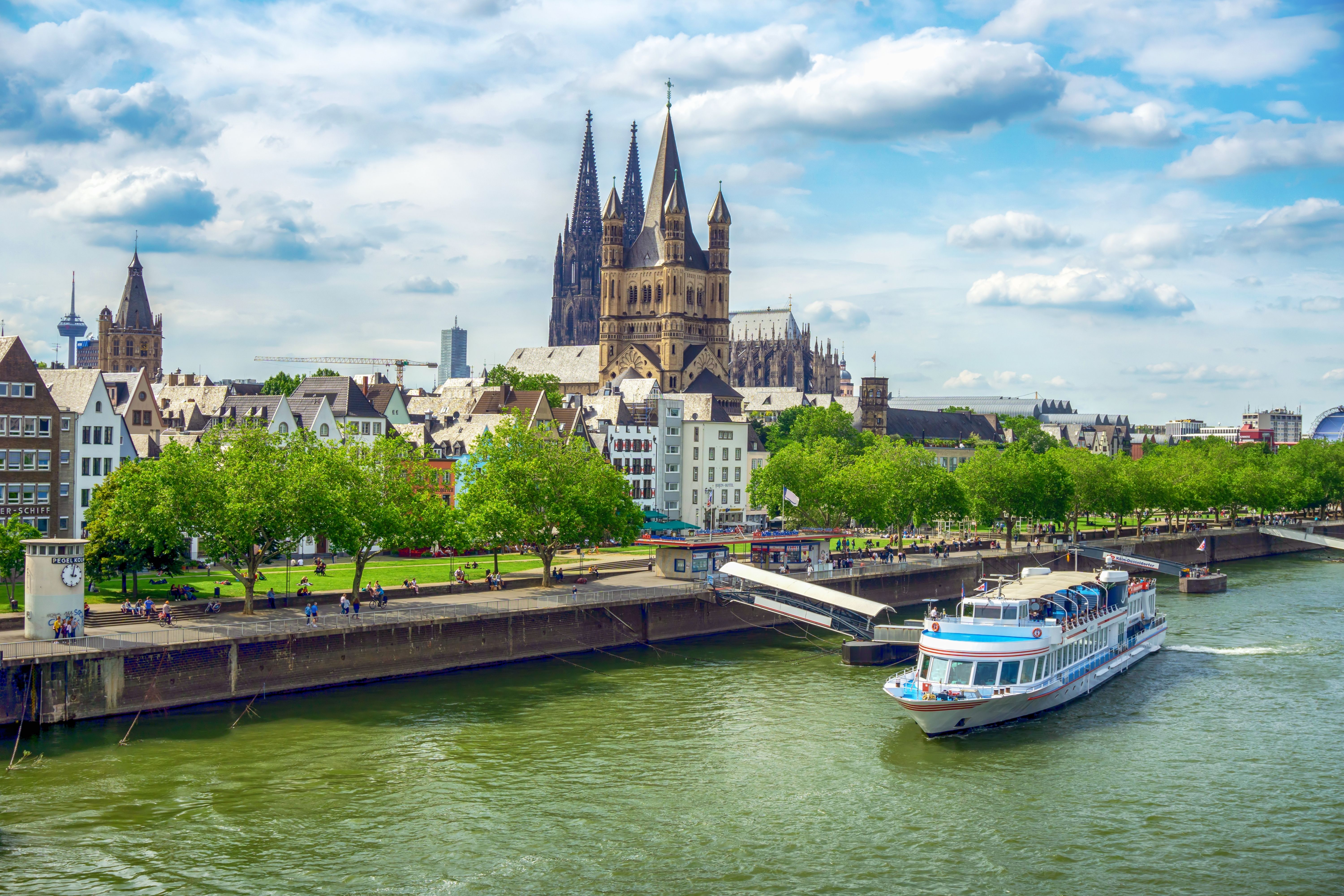 Cologne Cathedral and Rhine River cruise boat along Cologne waterfront in Germany under a bright blue sky with historic Old Town skyline.