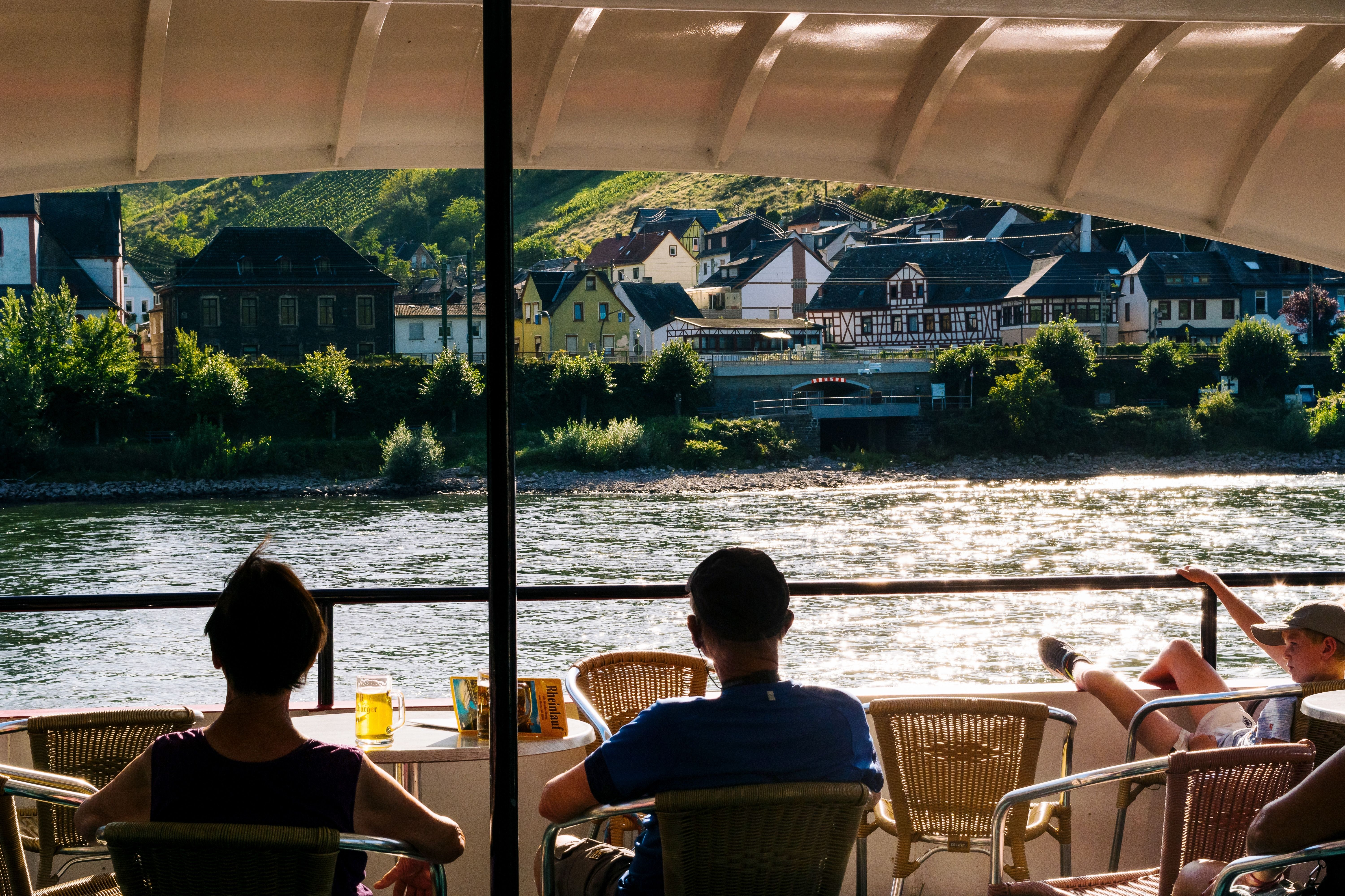 River cruise guests relaxing on open-air deck while sailing past a charming German Old Town village along the scenic Rhine River at sunset.