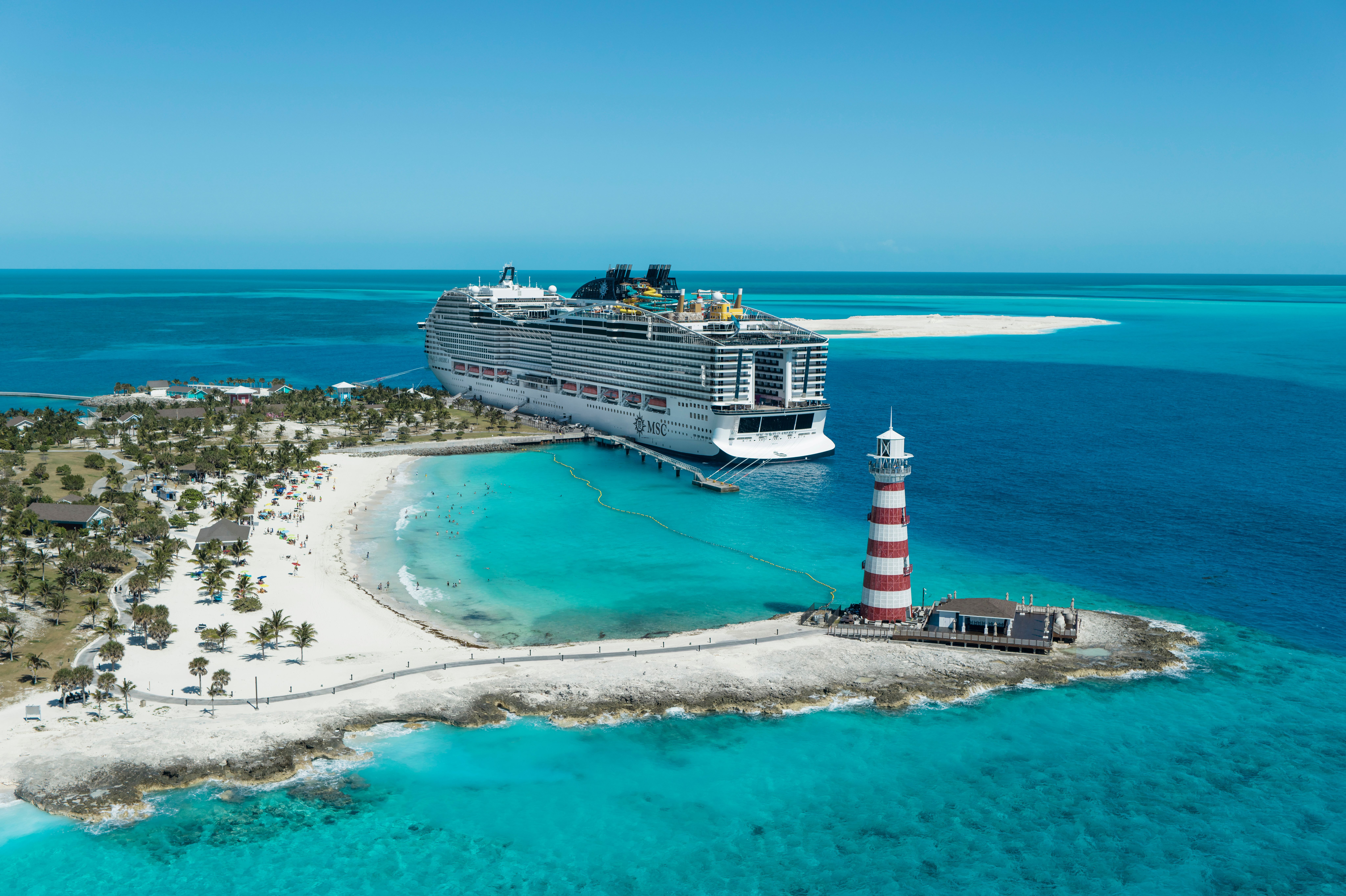 MSC cruise ship docked at Ocean Cay MSC Marine Reserve in the Bahamas, beside a white-sand beach, striped lighthouse and turquoise Caribbean waters.