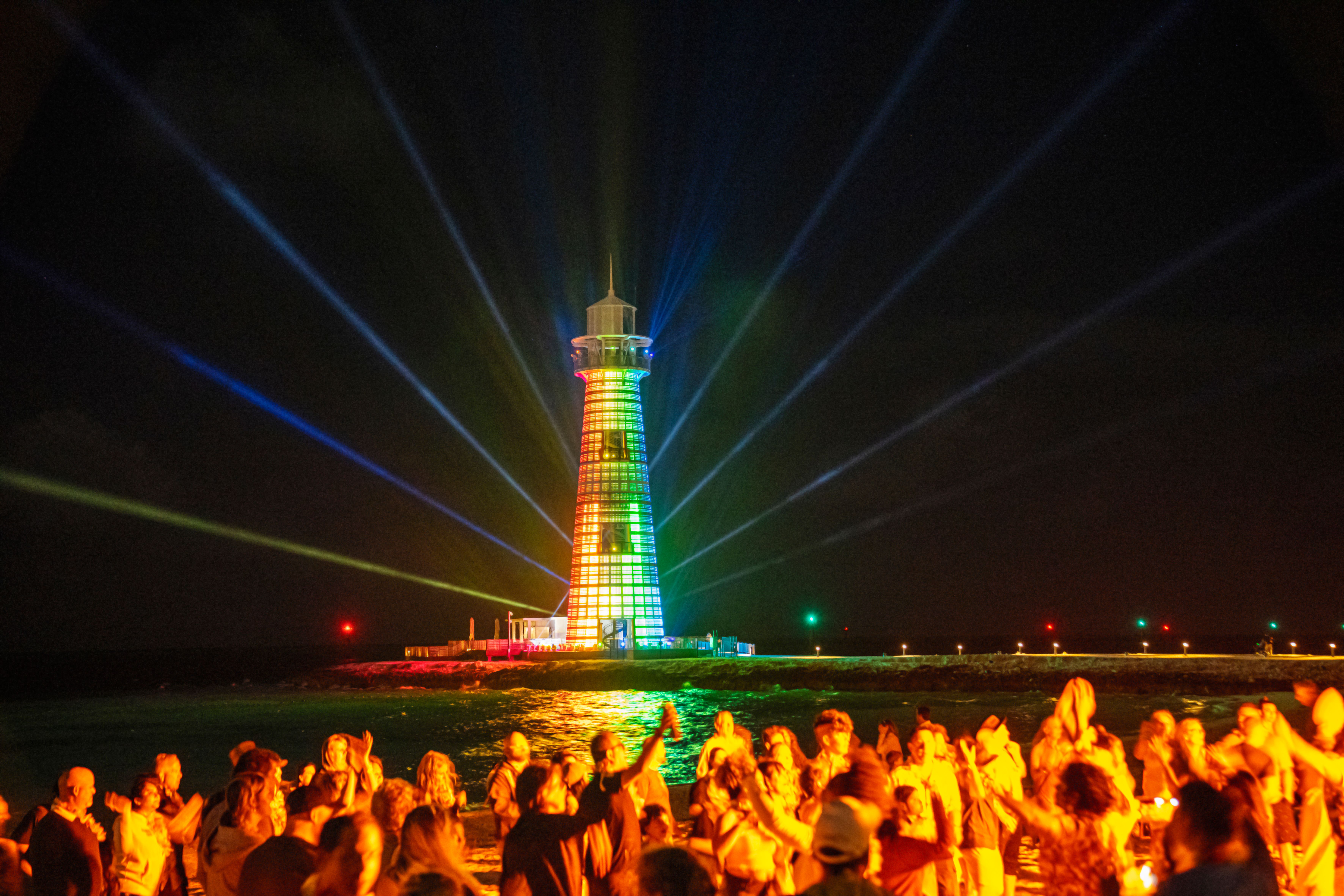 Colorful LED lighthouse show with rainbow lights and sweeping beams illuminating a waterfront crowd during a nighttime cruise destination event.