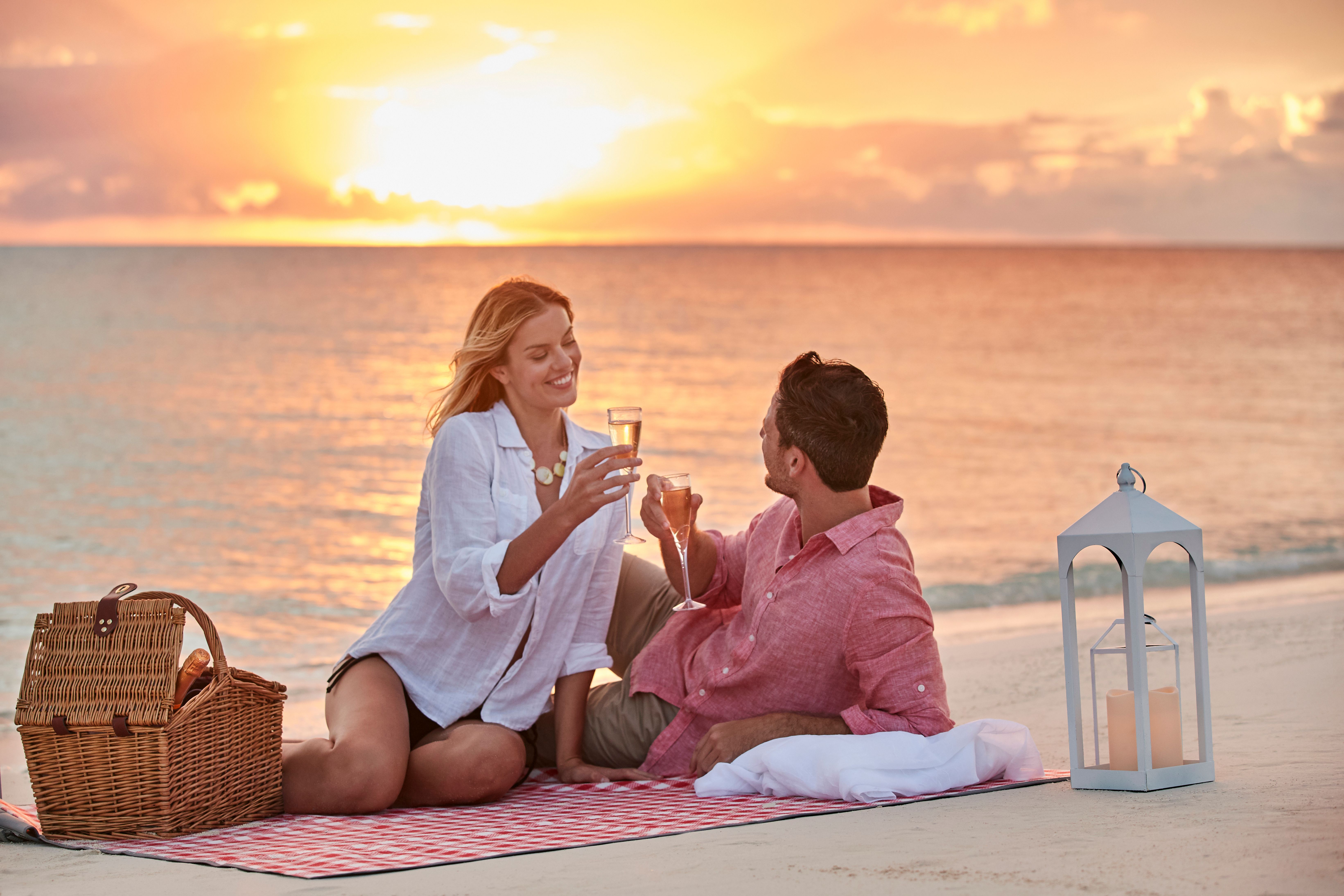 Couple enjoying a romantic sunset beach picnic with champagne, a lantern and ocean views during a cruise vacation night experience.