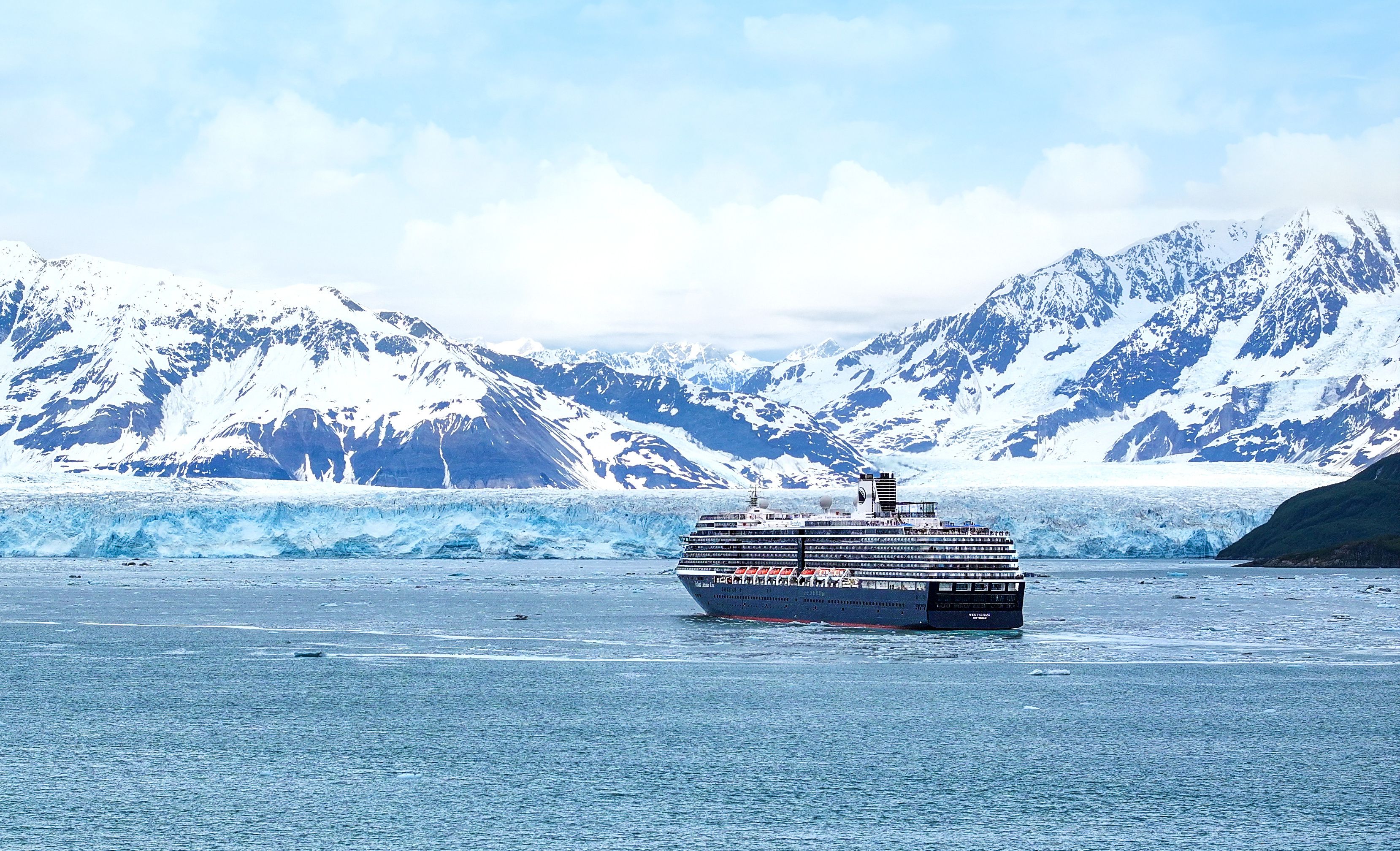 Holland America Line cruise ship sailing past a massive Alaska glacier and snow-covered mountains on a scenic Inside Passage cruise.