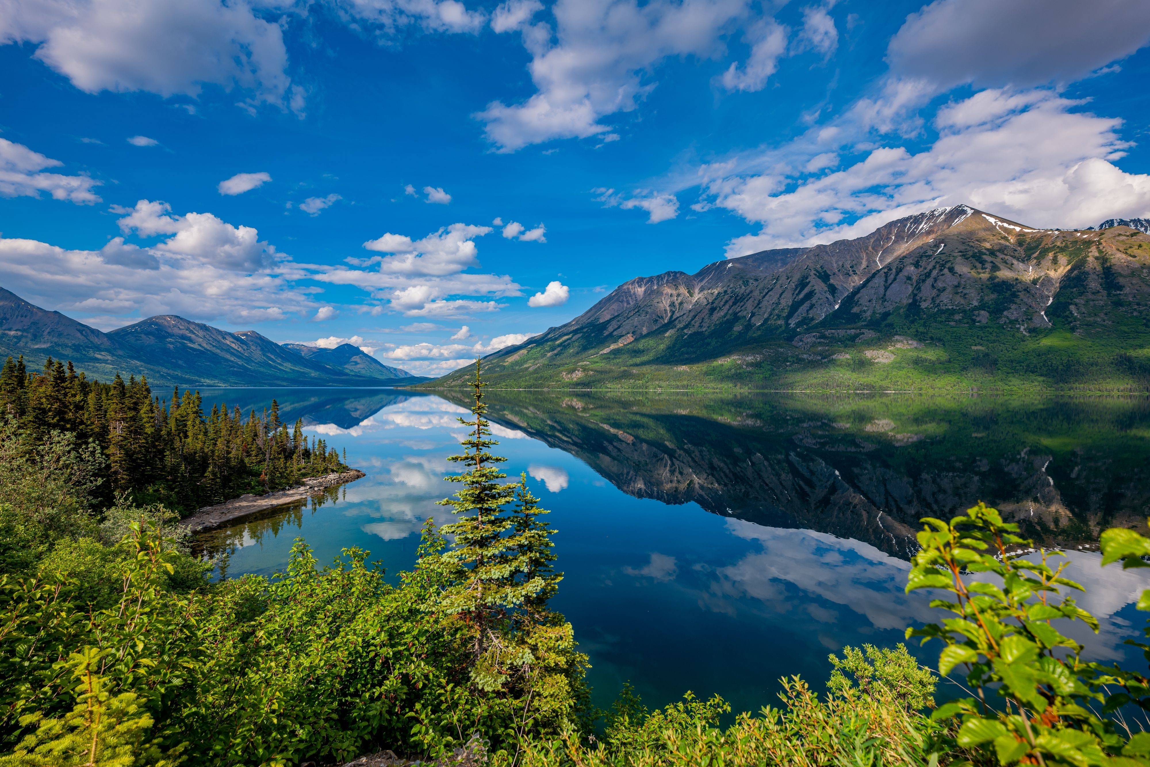 Scenic Skagway, Alaska landscape with a tranquil mountain lake, evergreen forest and rugged peaks reflected beneath a vivid blue sky.