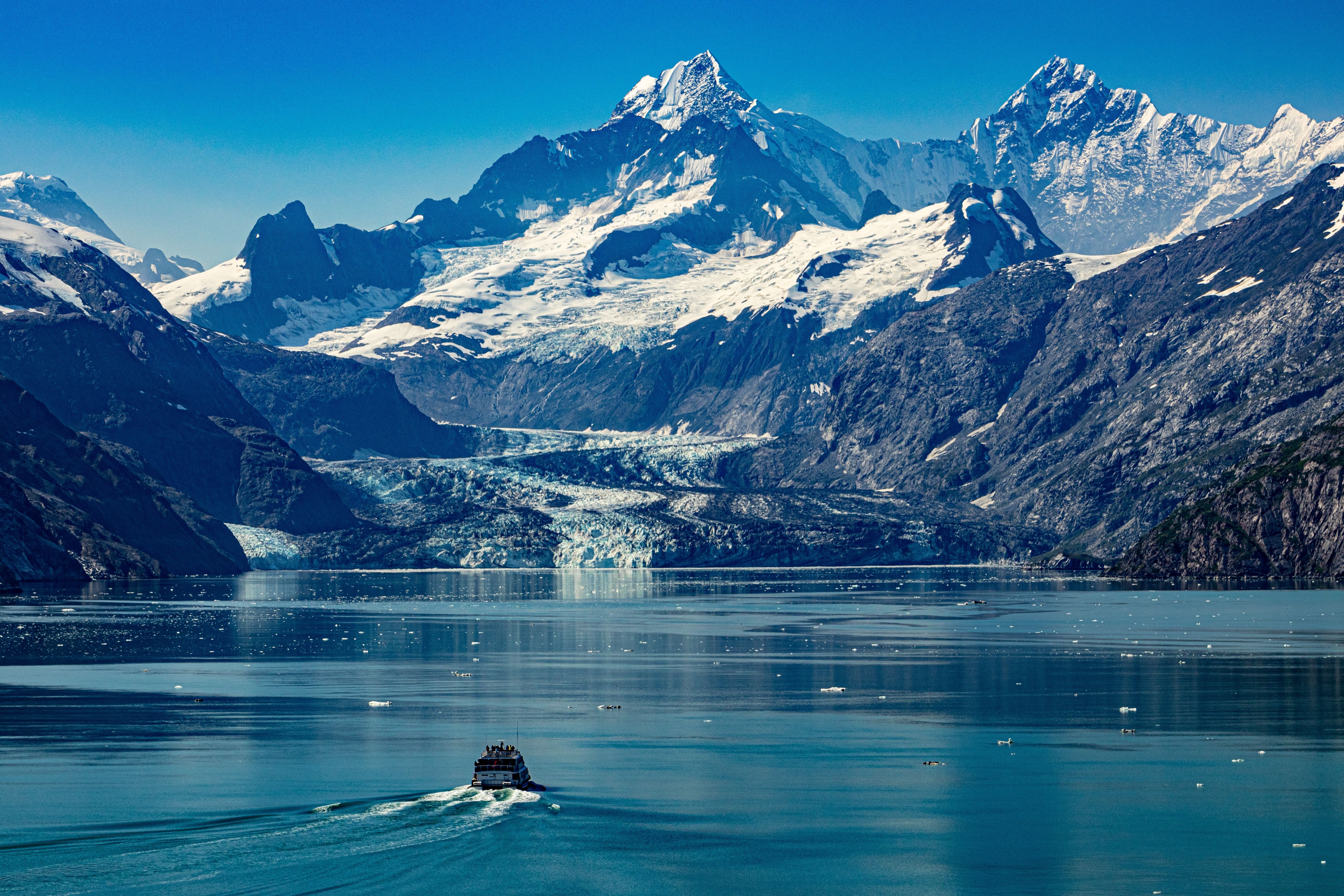 Glacier Bay National Park, Alaska, with a tour boat cruising icy blue waters toward a vast glacier framed by rugged snowcapped mountains.