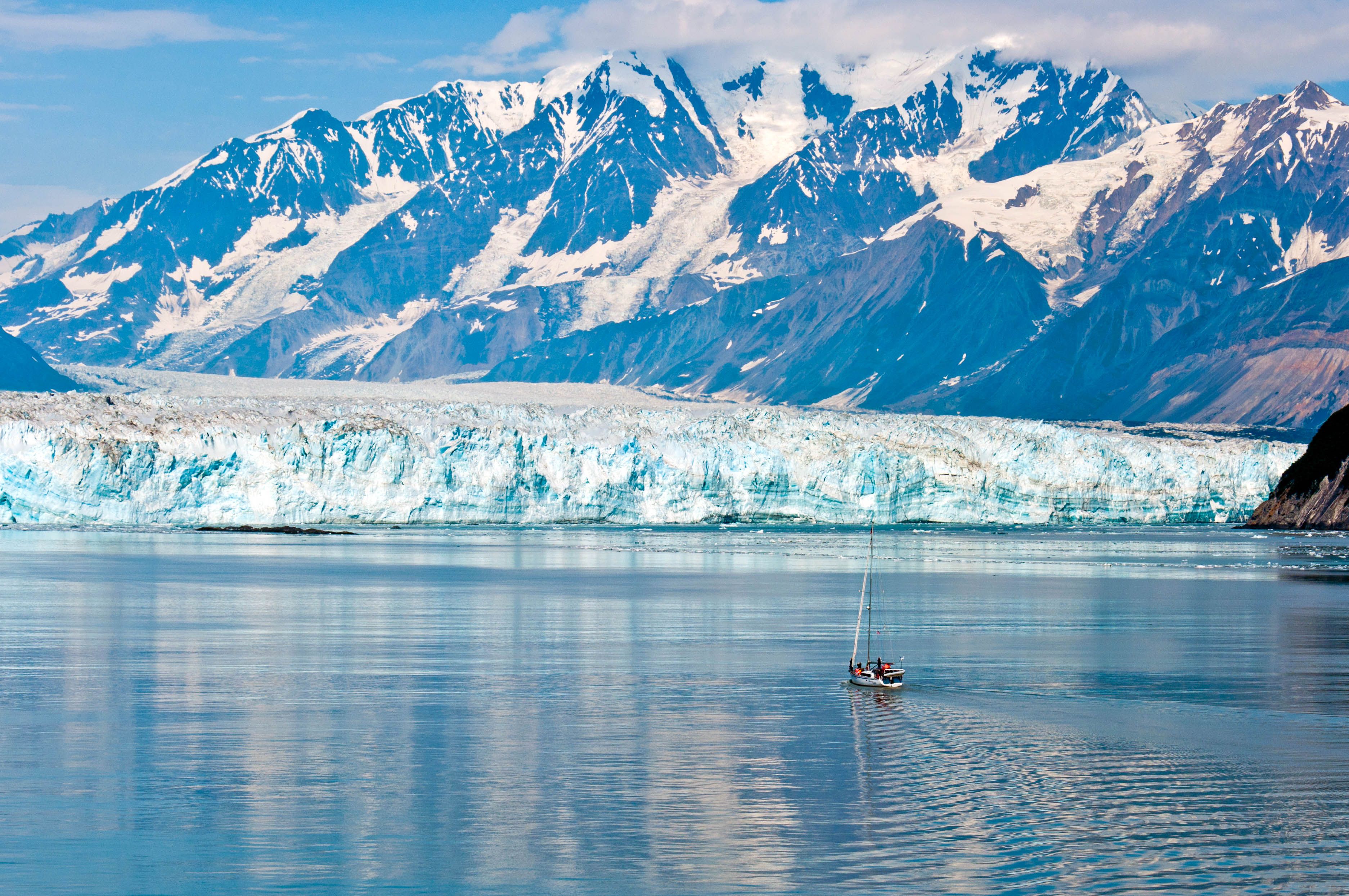 Hubbard Glacier in Alaska with a sailboat on calm blue water, framed by the massive glacier face and snow-covered coastal mountains.