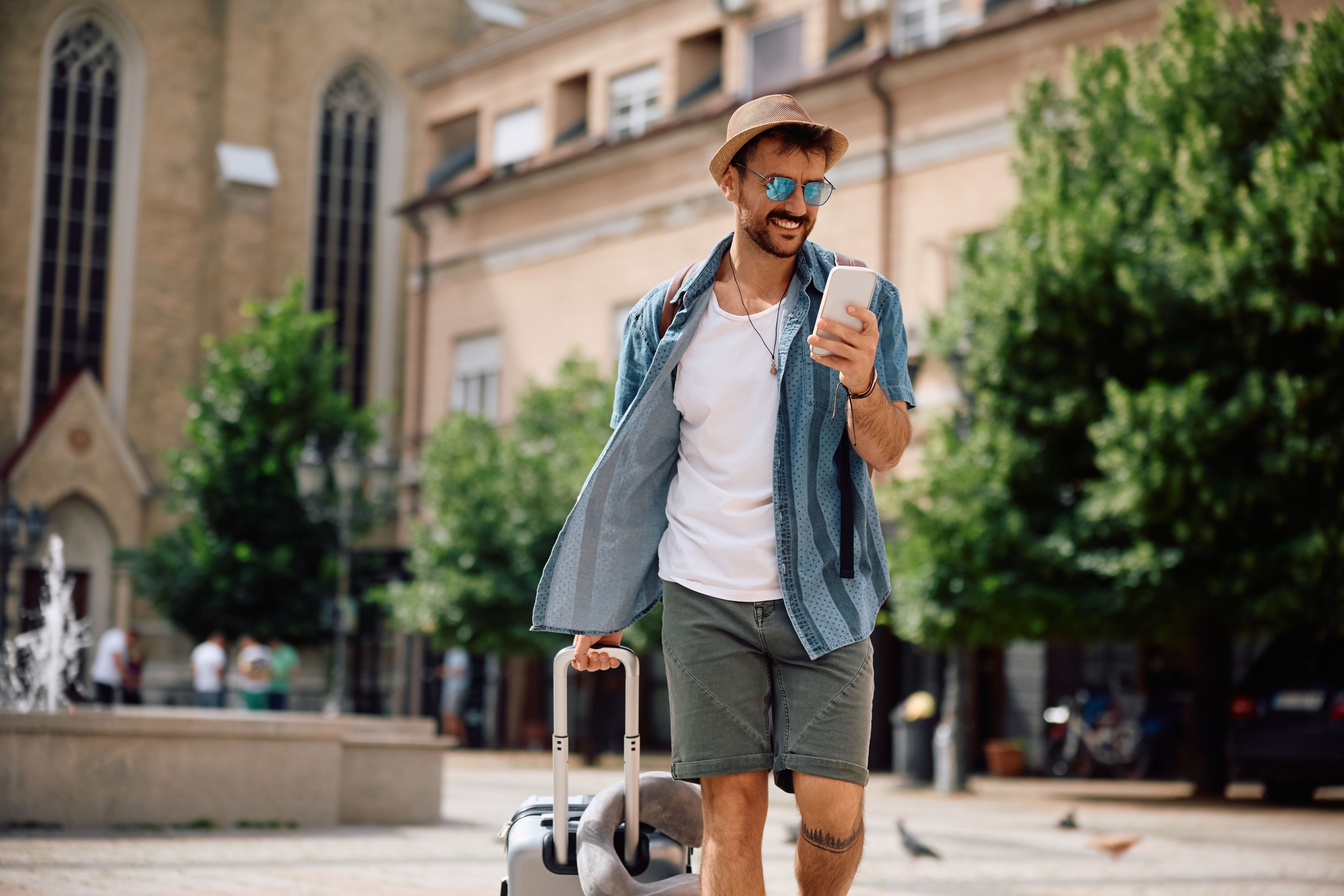 Smiling male traveler wearing sunglasses and a sunhat checks his smartphone while pulling a suitcase through a historic city square, illustrating travel safety tips.
