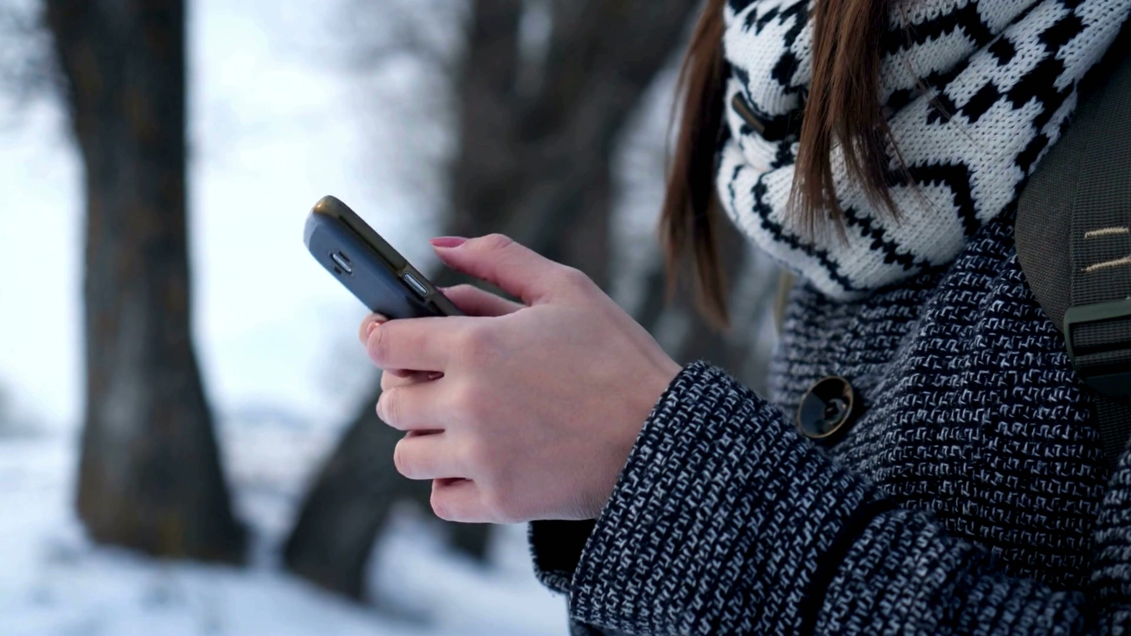 Person in a winter coat and patterned scarf using a smartphone outdoors, with snow-covered trees in the background.