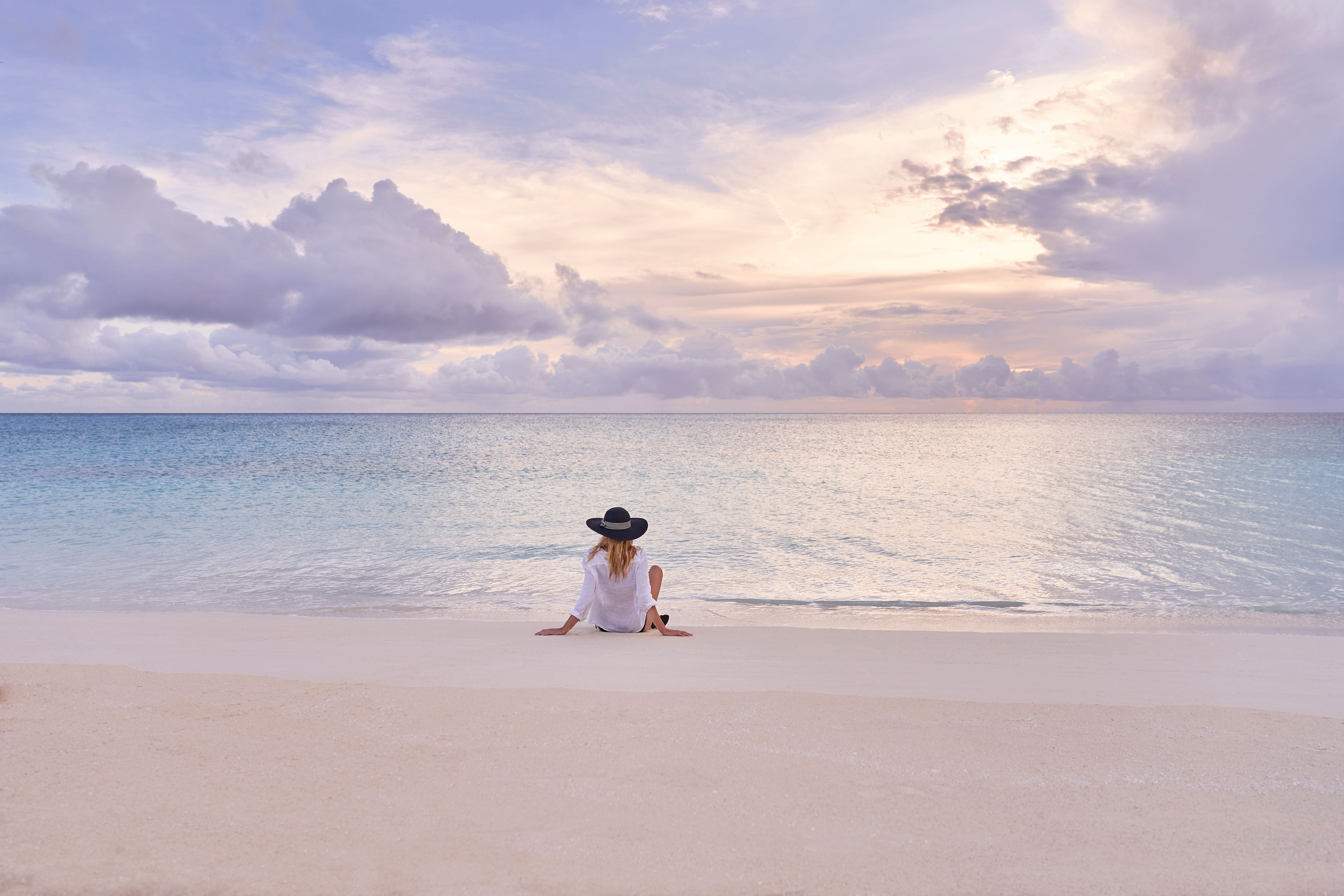 Woman relaxing on a pristine white sand beach at MSC Cruises' Ocean Cay Marine Reserve, overlooking calm turquoise Caribbean waters beneath a pastel sunset sky.